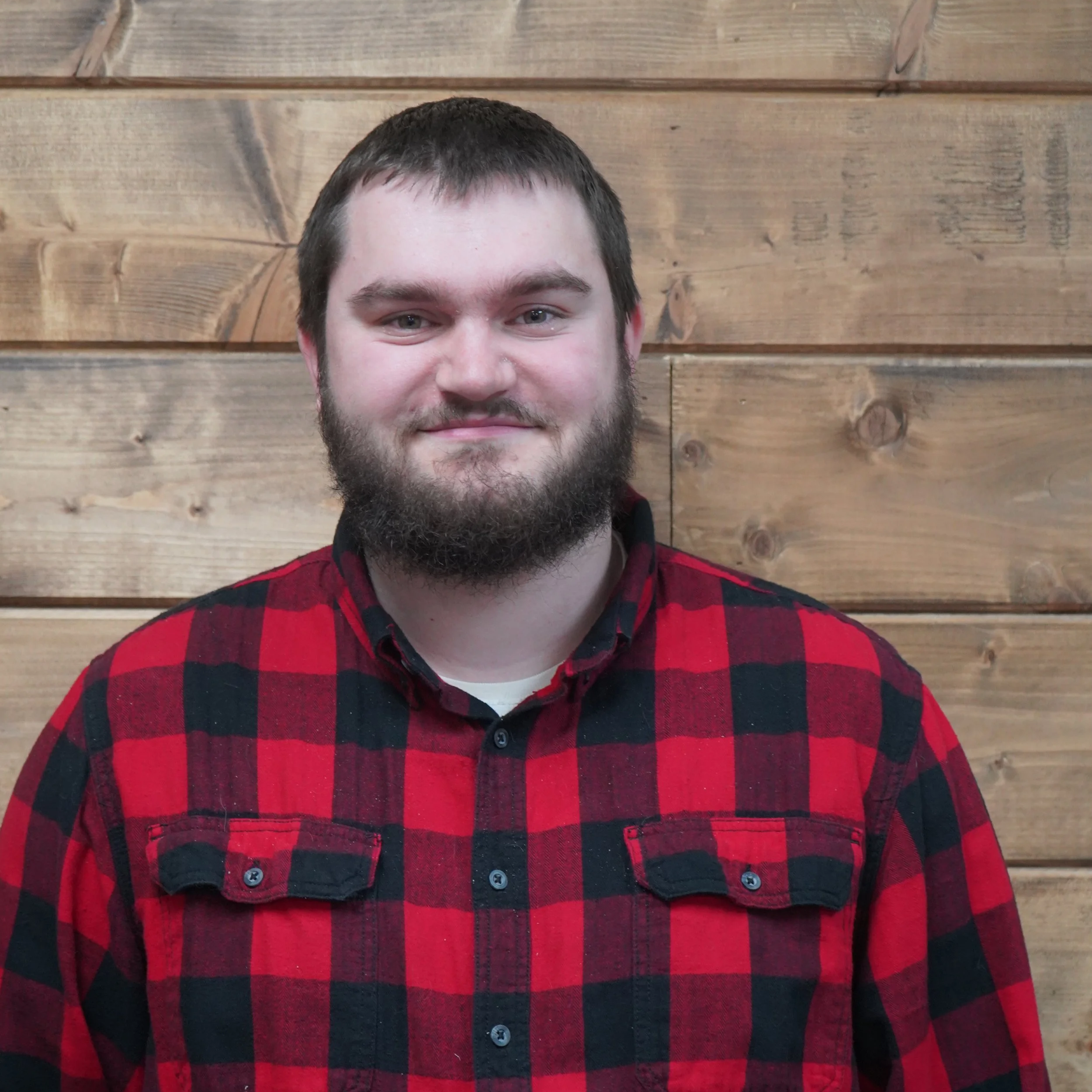 A man with a beard and short brown hair smiling, wearing a red and black plaid shirt, standing in front of a wooden wall.