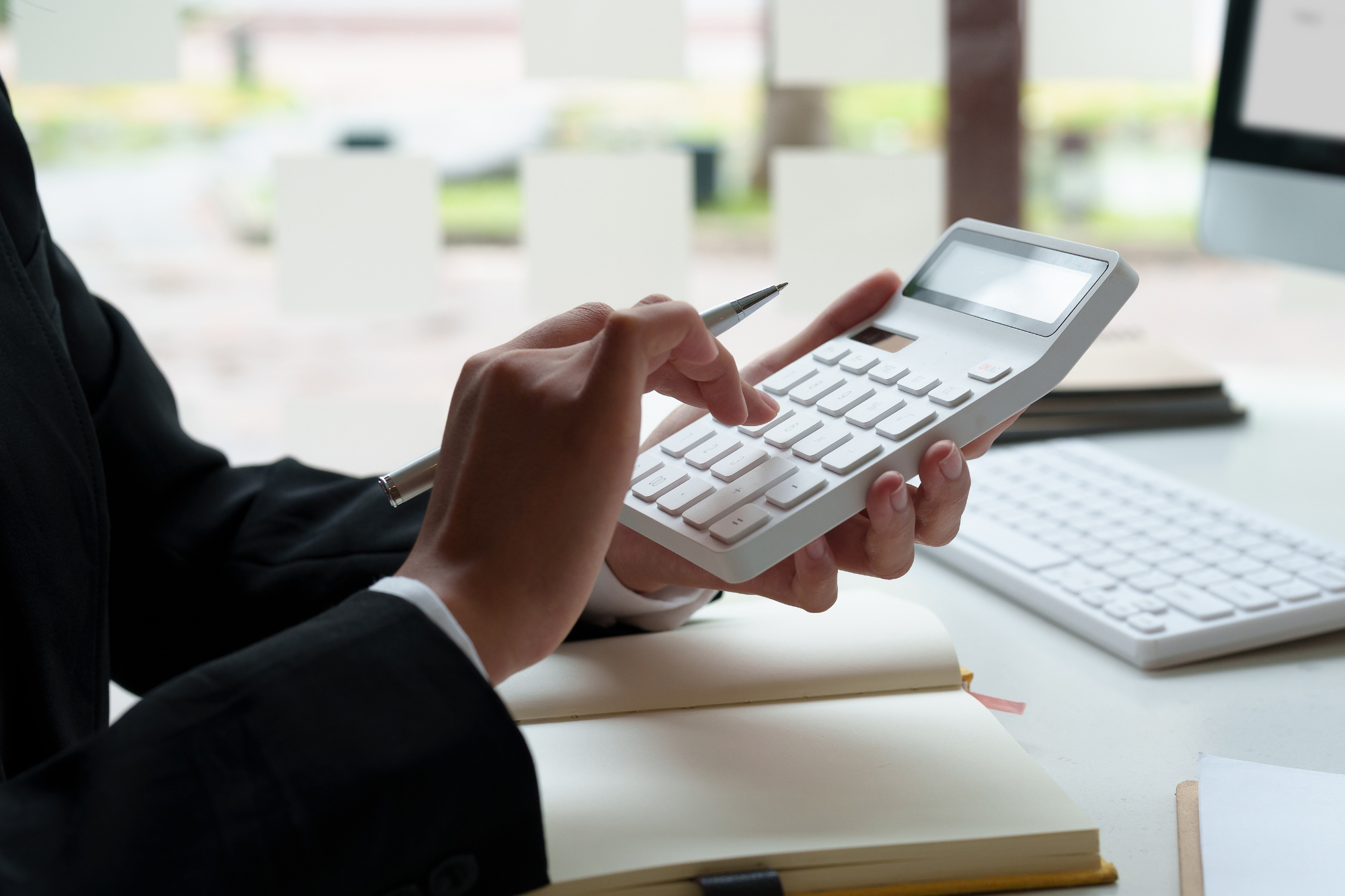 Personne utilisant une calculatrice tout en tenant un stylo et prenant des notes dans un cahier, avec un clavier d'ordinateur à proximité.