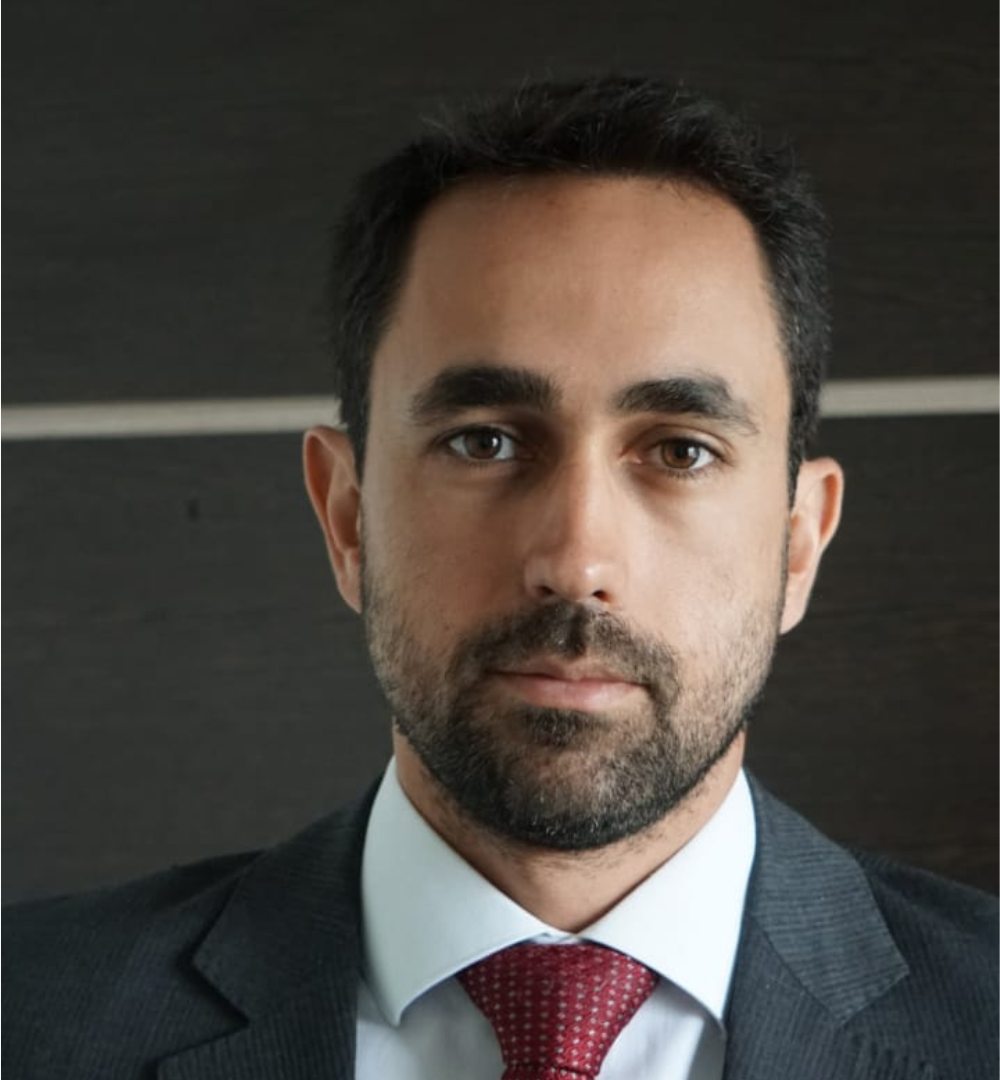 Man with a beard wearing a suit and red tie against a dark background.