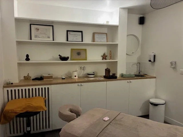 Une salle de soins avec un lit, des étagères blanches avec des diplômes et des décorations, et un espace de lavabo avec un évier.