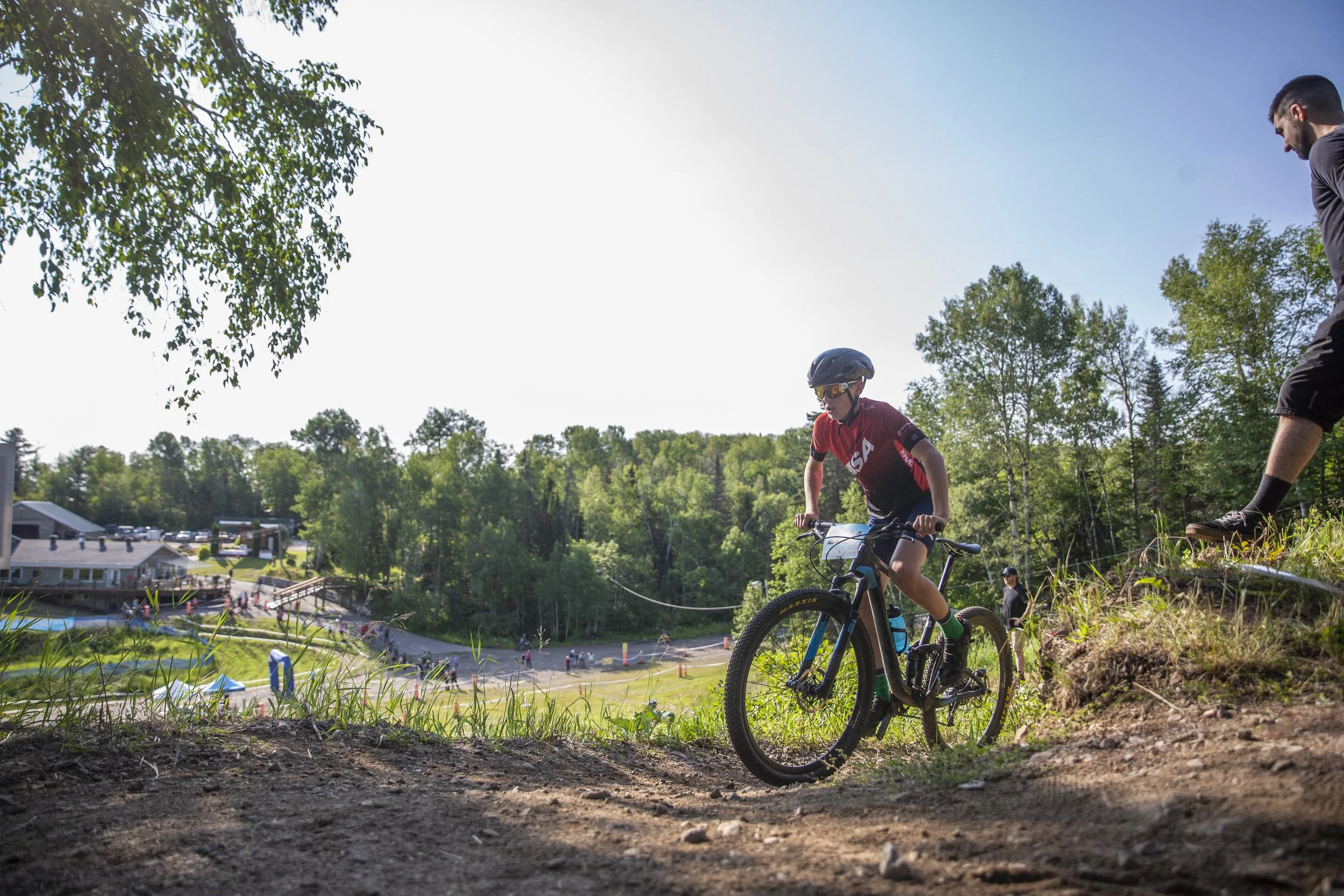 Un jeune garçon en casque et chemise de sport rouge à vélo, en descente sur un sentier de montagne, dans un environnement naturel avec des arbres et un bâtiment au fond.