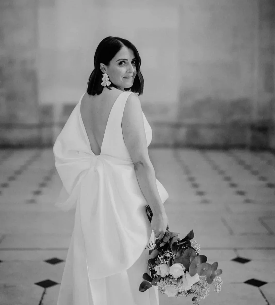 A woman in a white dress holding a bouquet in a large, elegant hall with a checkered floor, looking over her shoulder and smiling.
