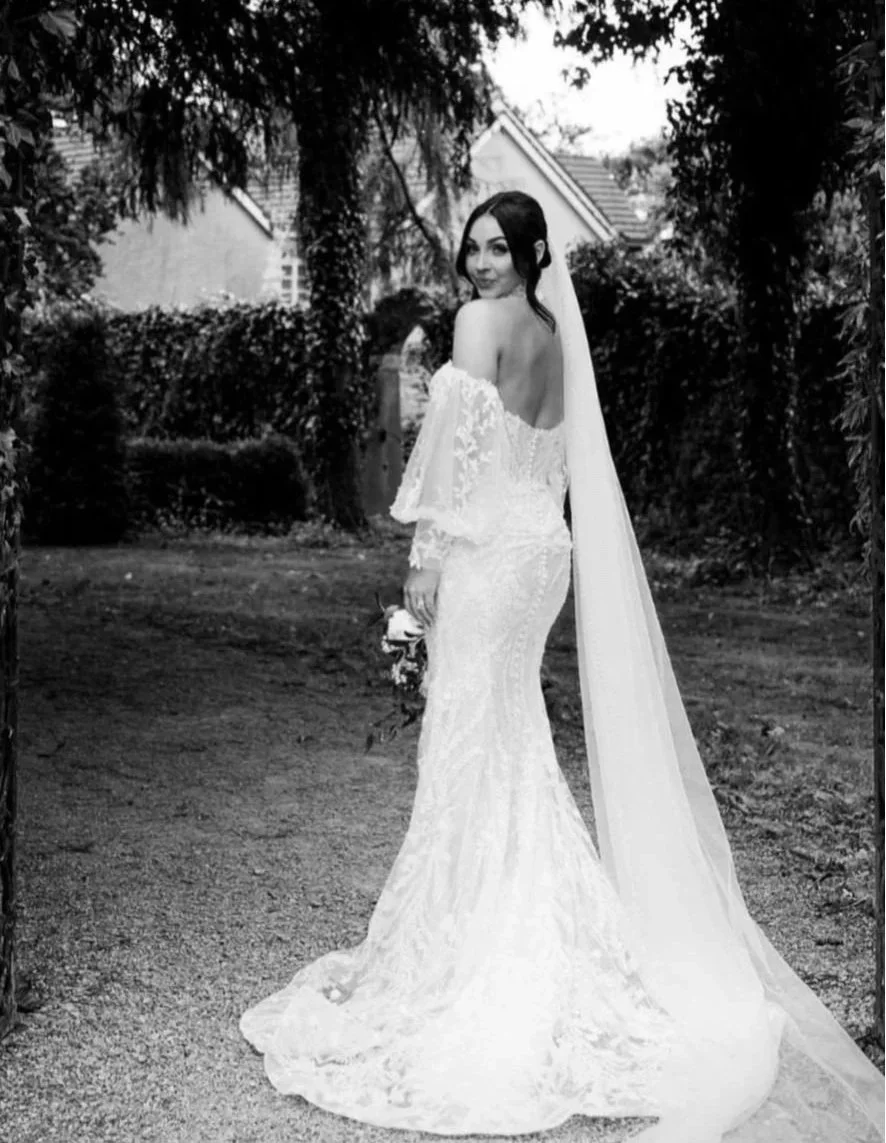 A bride in a white wedding dress holding a bouquet, standing outdoors among trees and bushes, with a house in the background.