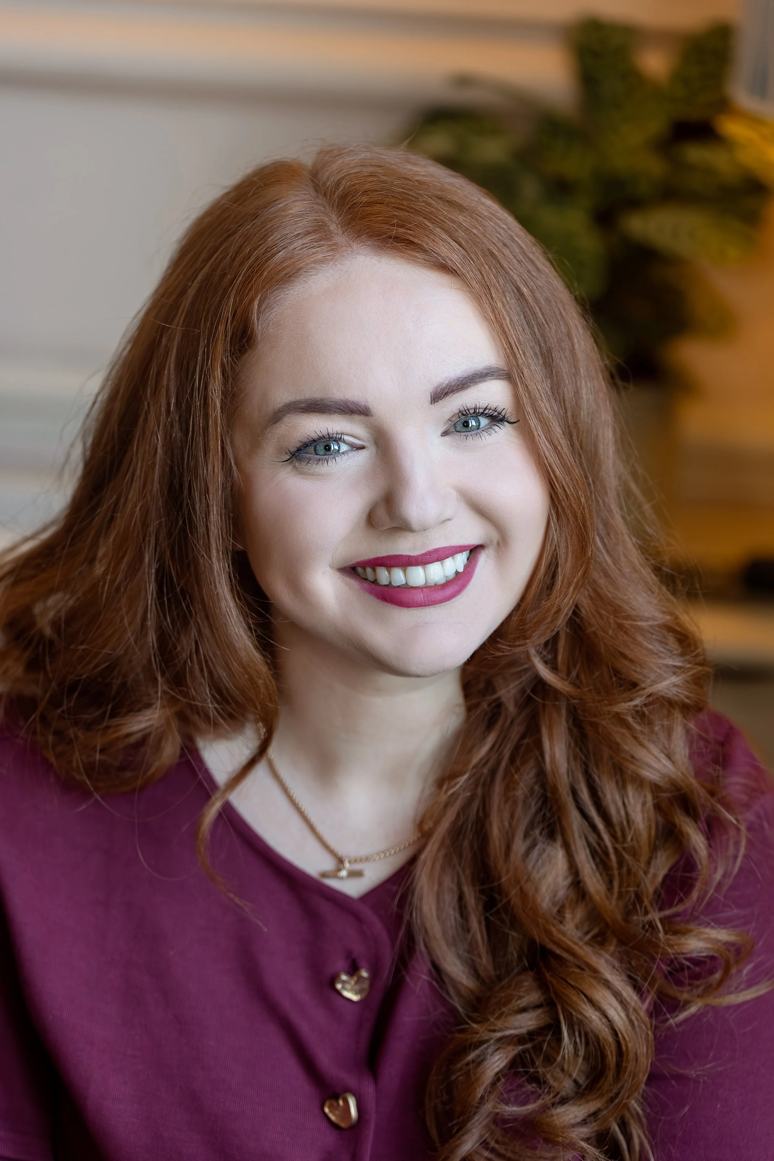 Close-up photo of a woman with long, wavy red hair, blue eyes, and bright red lipstick, smiling and wearing a shimmering black and gold striped top, outdoors with blurred background.
