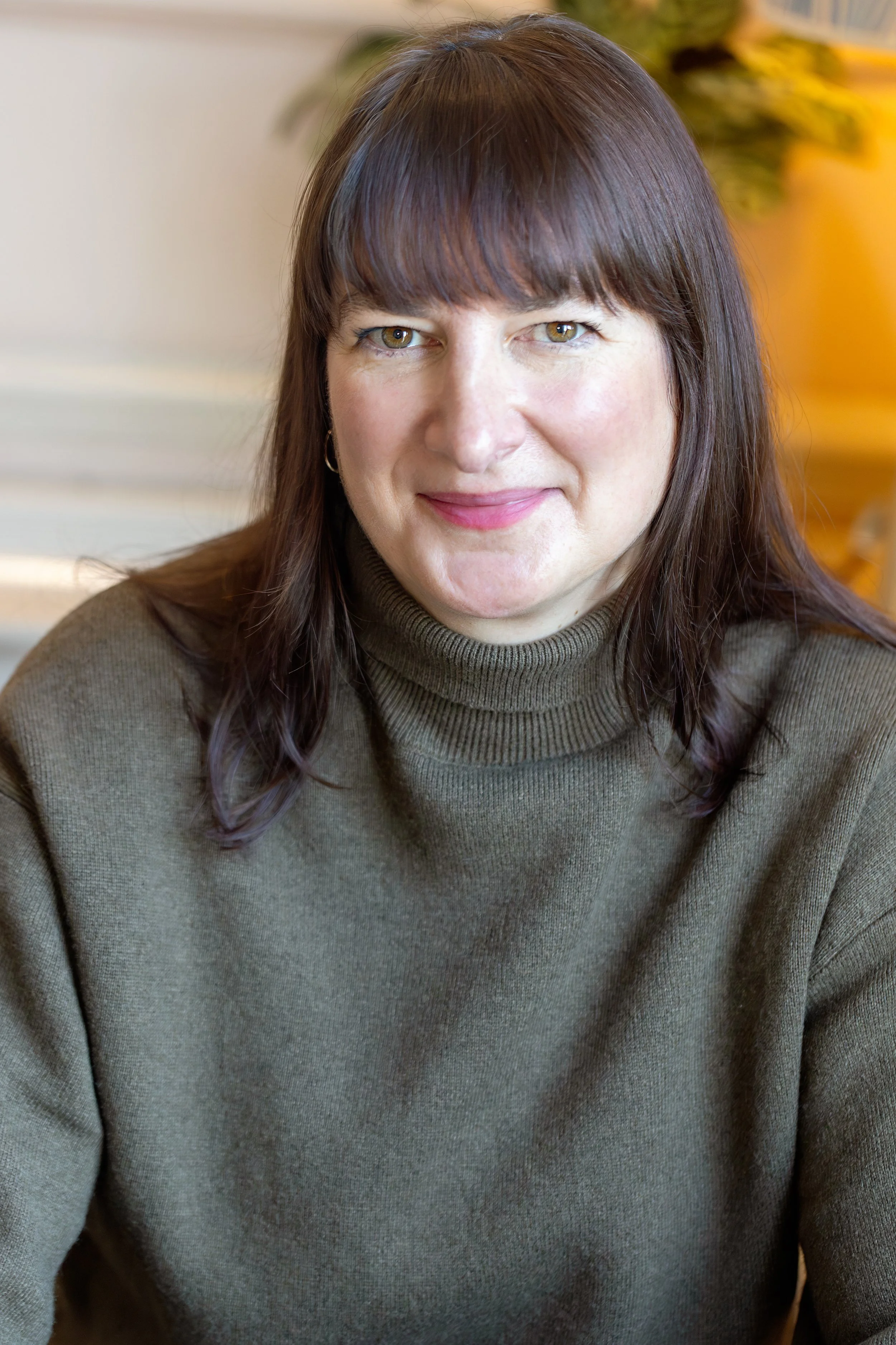 A woman with brown hair and bangs, wearing a green turtleneck sweater, smiling at the camera indoors with a blurred background including a plant.