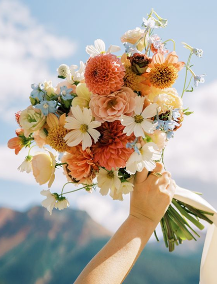 Buquê de flores coloridas sendo segurado por uma mão com fundo de céu azul e montanhas.