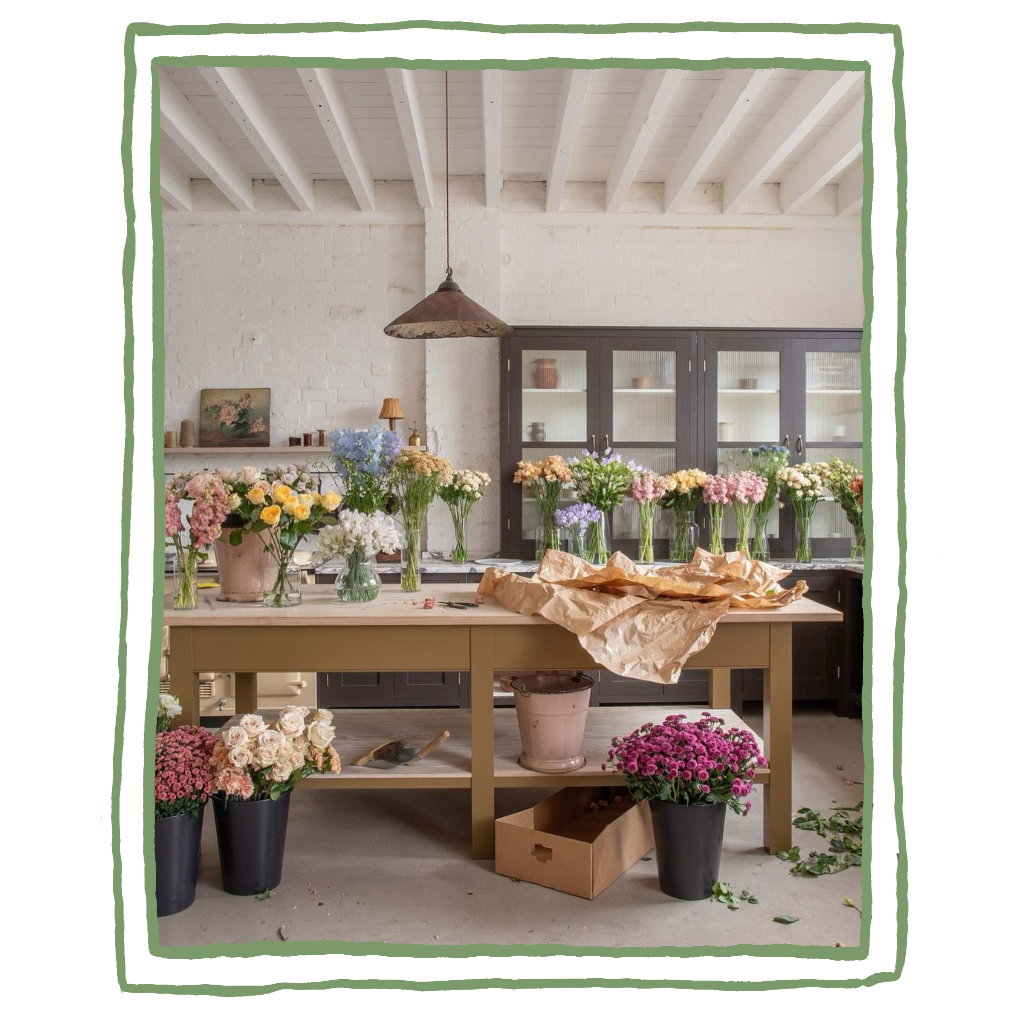 Oficina de flores com bancadas cheias de arranjos florais, vasos de vidro, flores coloridas em recipientes e ferramentas de jardinagem em um ambiente rústico e iluminado.