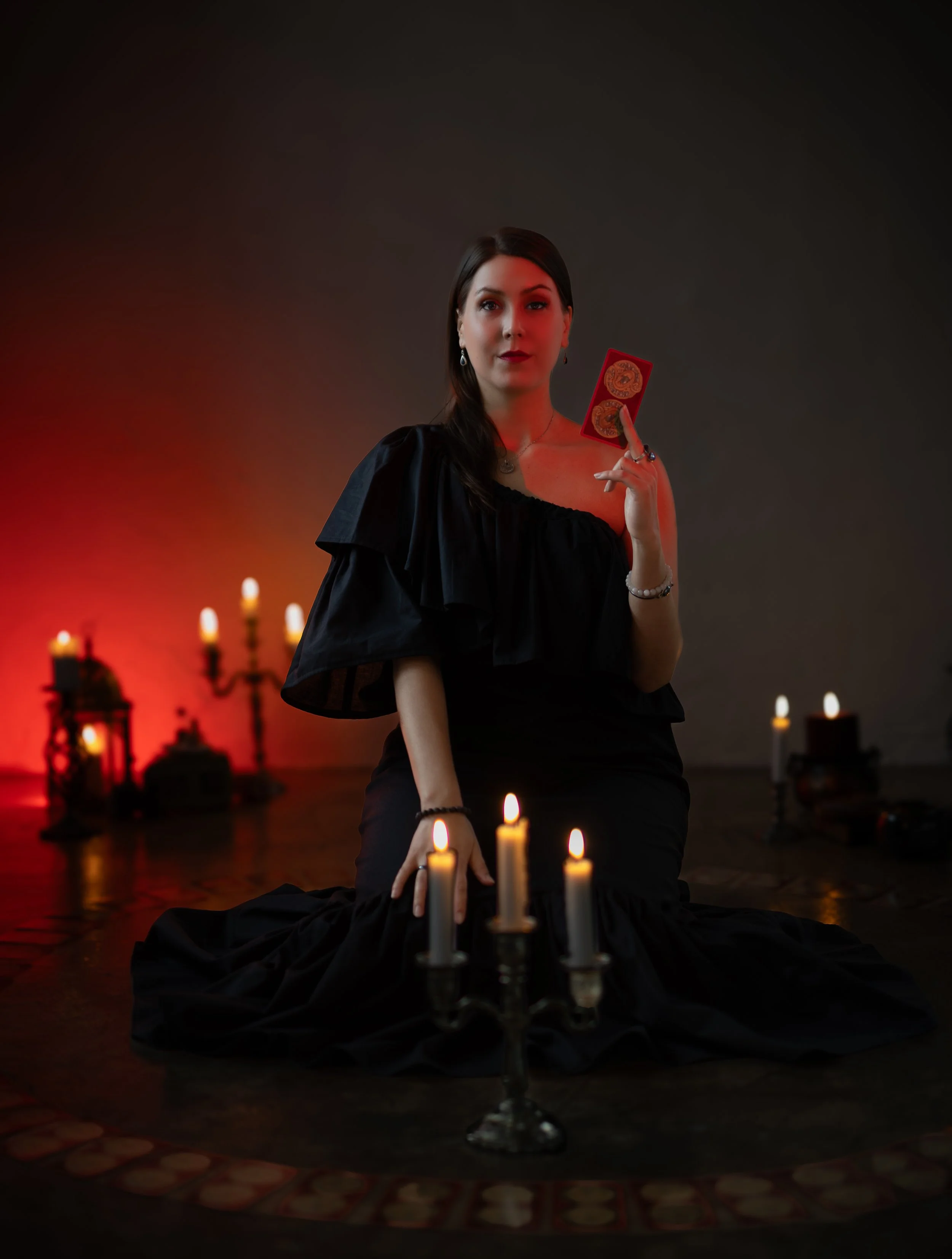 Meeri Toivonen istuu tarot-korttien keskellä punaisessa valaistuksessa. Kynttilöitä edessä ja takana. Musta mekko ja yksi tarot-kortti kädessä. Noidan kehä KokoTeatterissa maaliskuussa 2026.