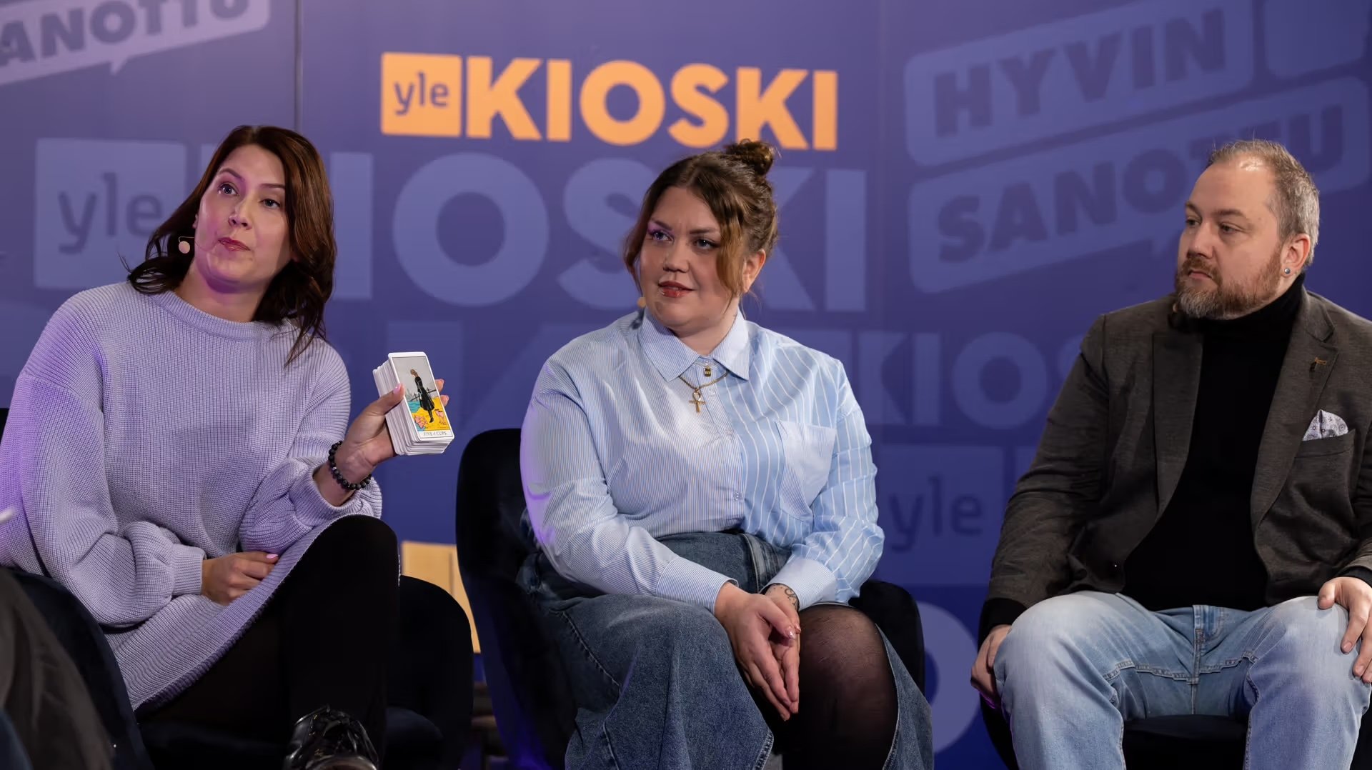 Kolme ihmistä istuu keskustelutilaisuudessa, Meeri Toivonen näyttää kortteja ja toinen nainen katsoo häntä, mies katsoo muualle. Taustalla teksti "Yle Kioski".