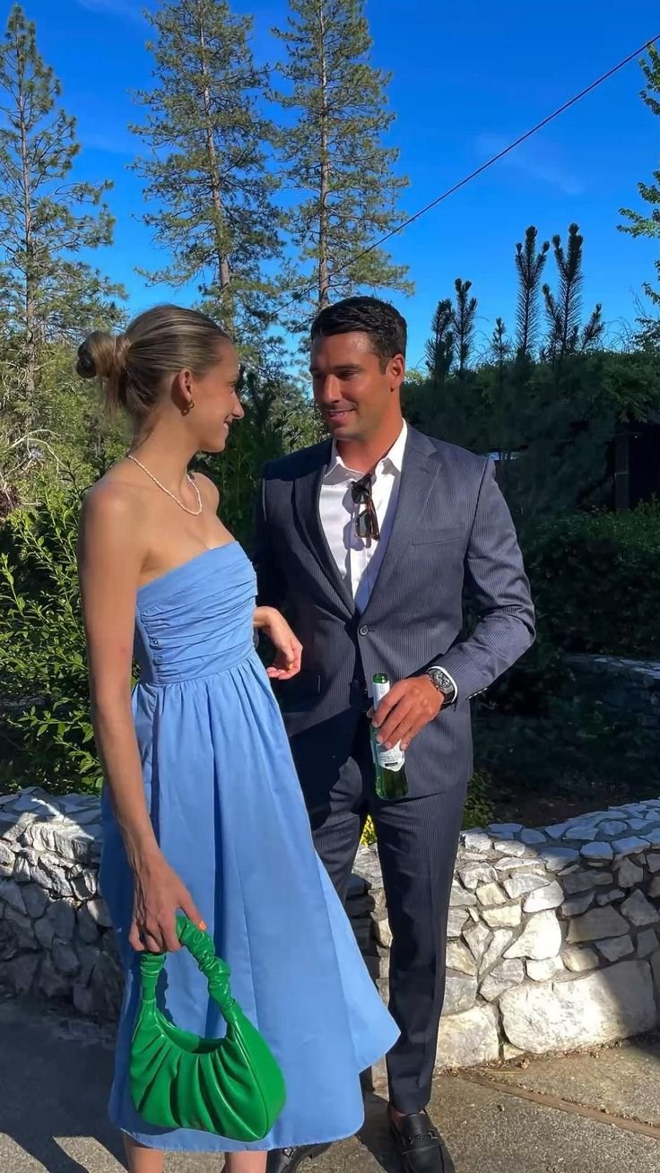 Woman in blue dress holding green bag, standing beside a man in a suit outdoors with trees and blue sky background.