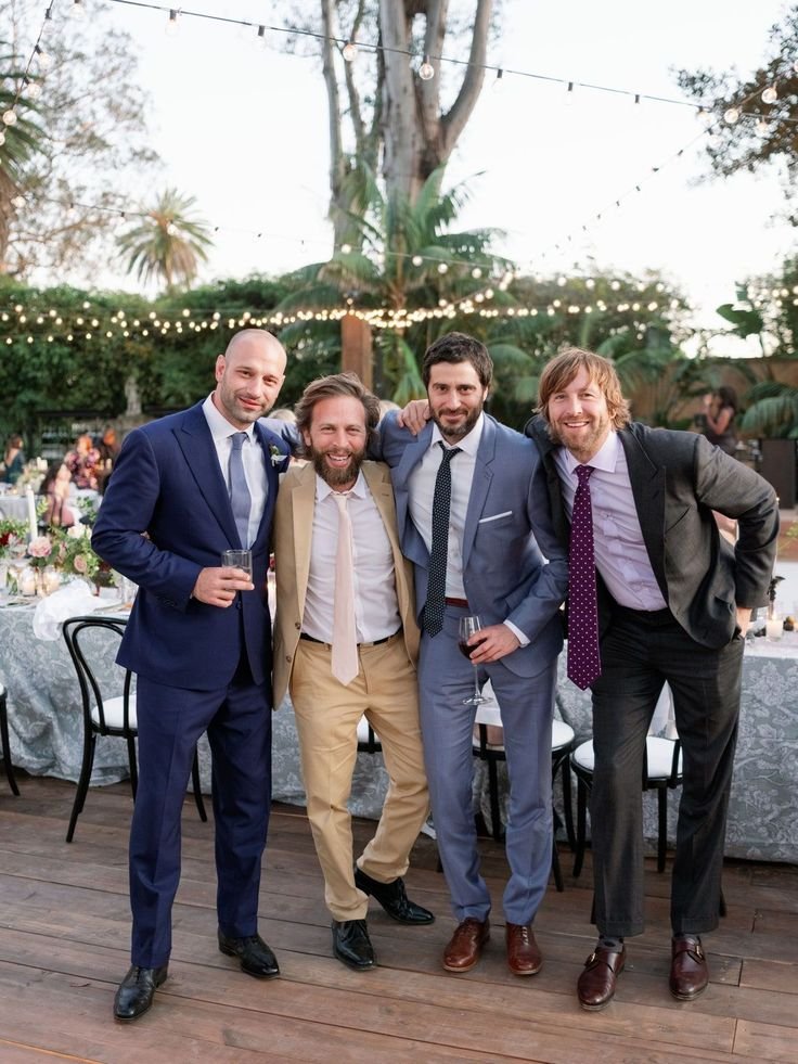 Group of four men in suits posing outdoors at a wedding reception with string lights and palm trees in the background.