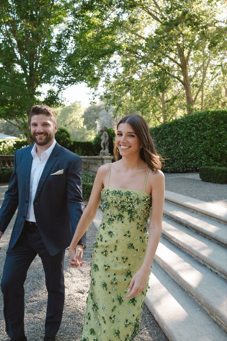 A couple holding hands, smiling, walking in a garden with greenery and a statue in the background.