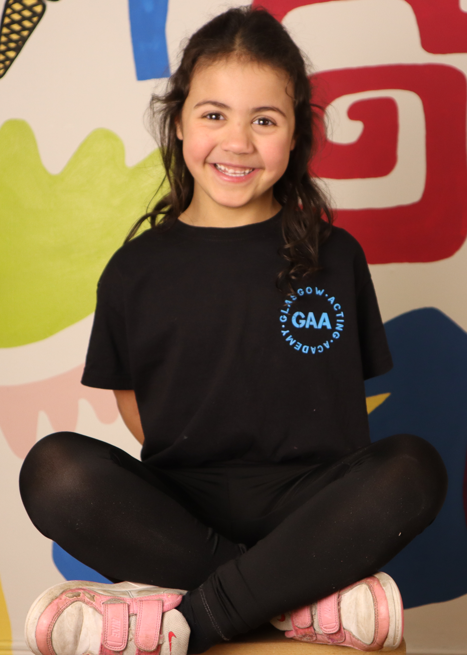 A young girl with brown hair sitting cross-legged, smiling at the camera. She is wearing a black T-shirt with a blue logo that says "GAA" and a background with colorful abstract shapes.