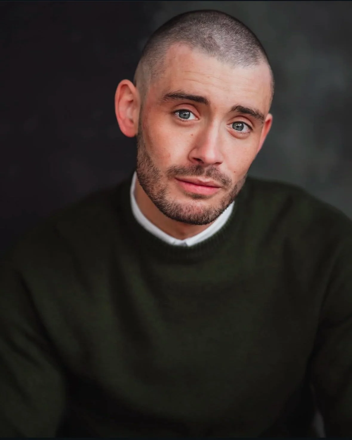 A young man with short hair and a beard looking at the camera, wearing a dark sweater with a white collar underneath, against a dark background.