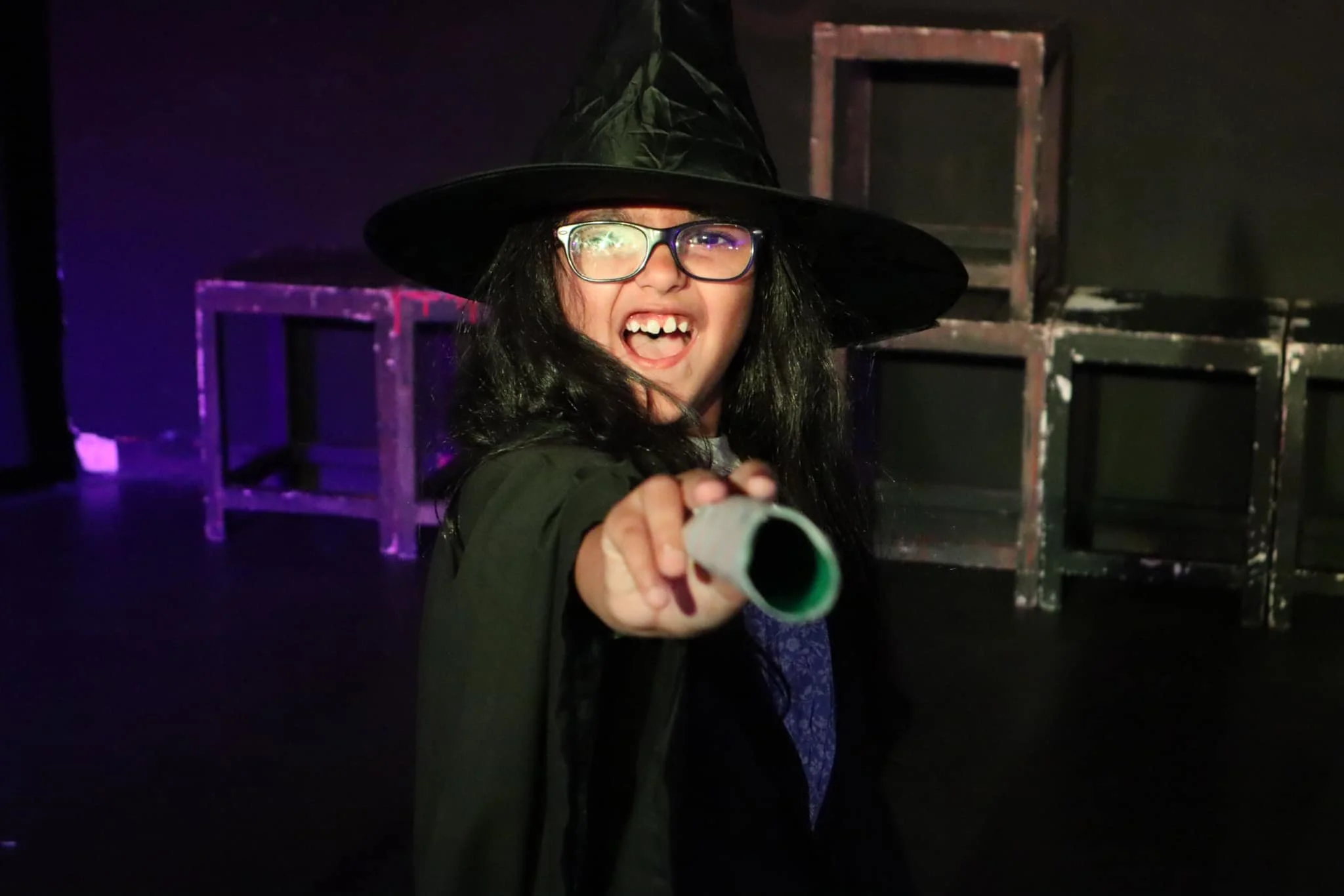A person dressed as a witch, wearing a black hat and glasses, pointing and smiling at the camera on a stage with empty chairs and purple lighting.