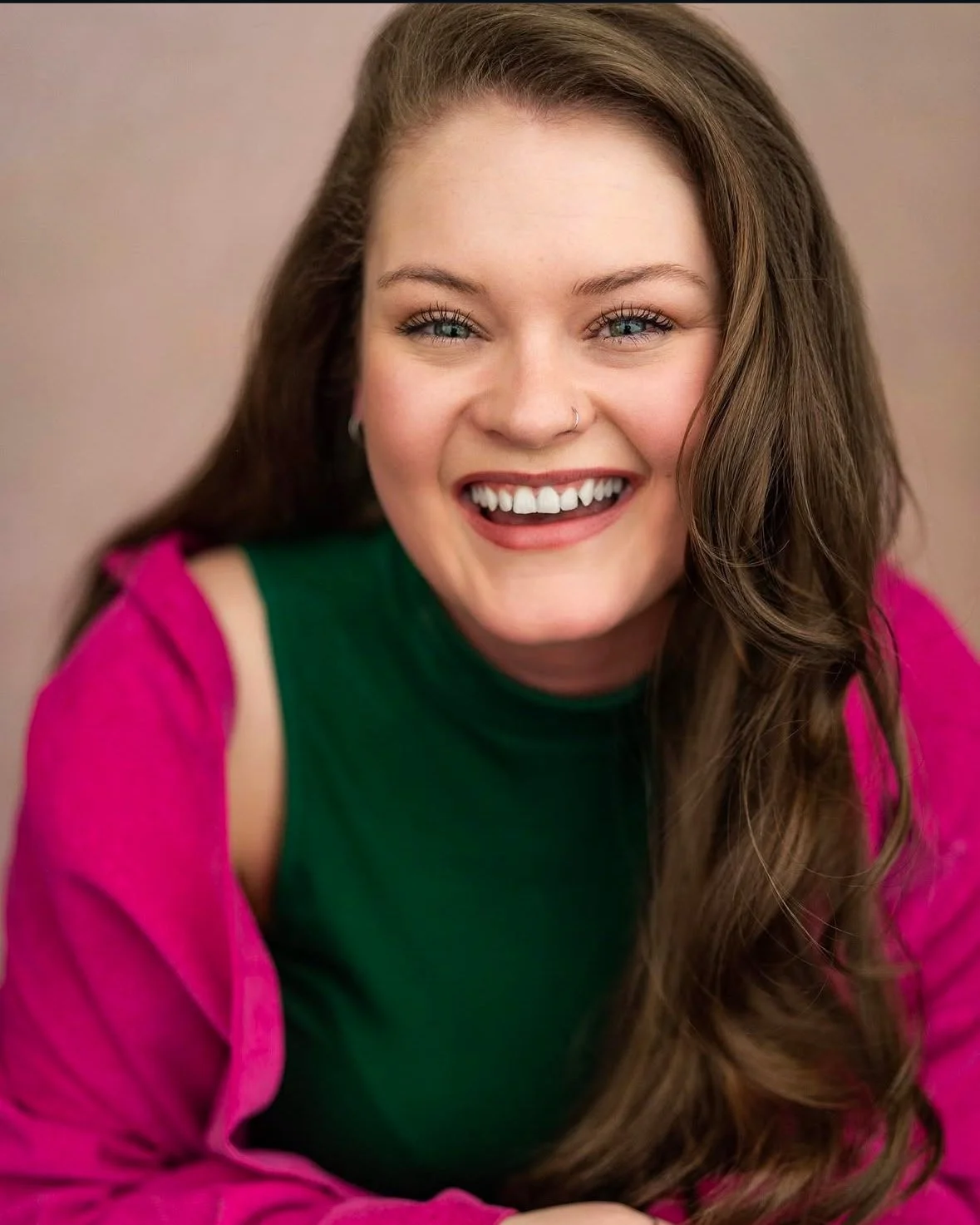A smiling young woman with long wavy hair, wearing a green top and a pink jacket, posing against a plain background.