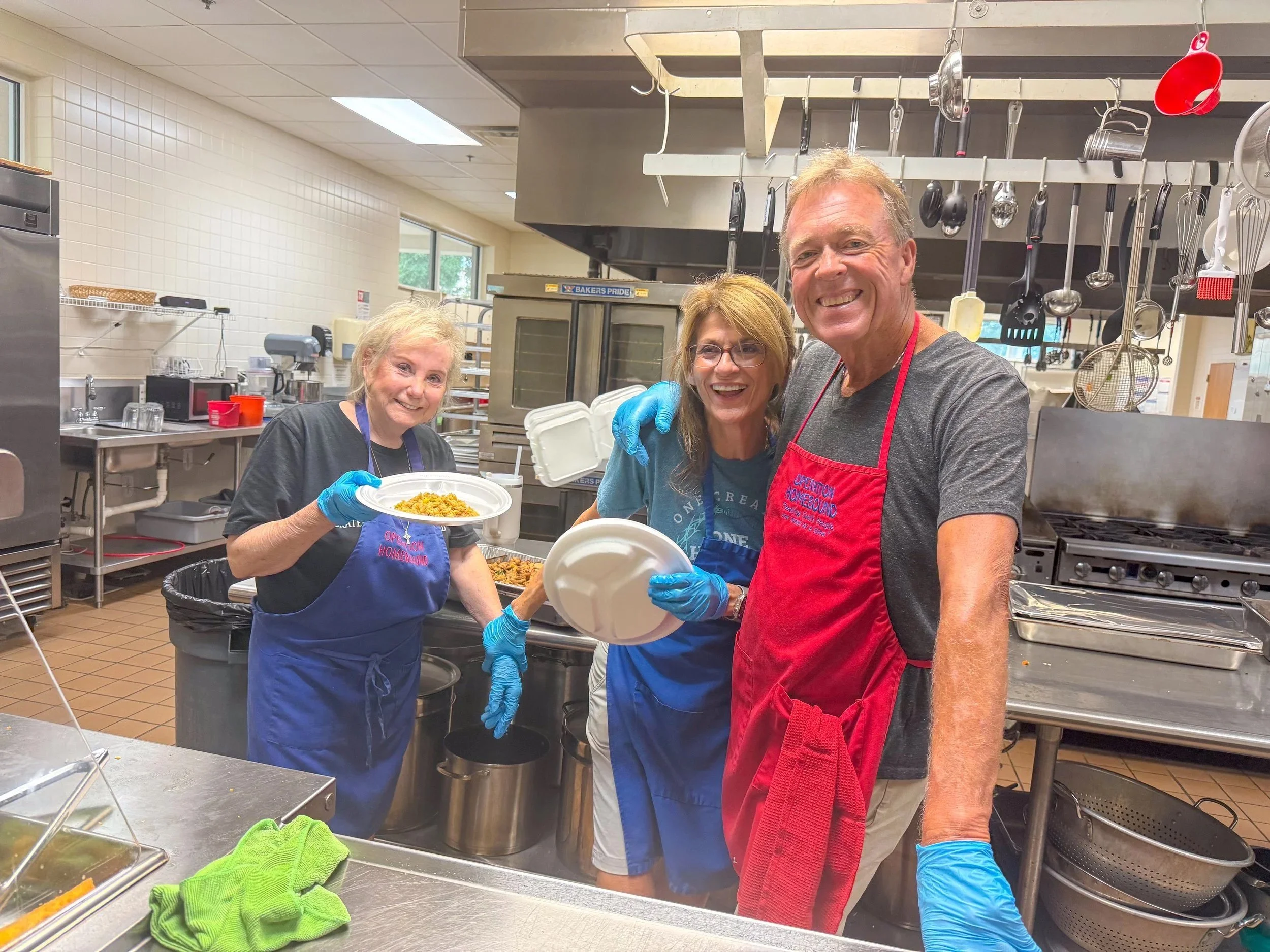 Three smiling volunteers in aprons and gloves serving meals in the church kitchen