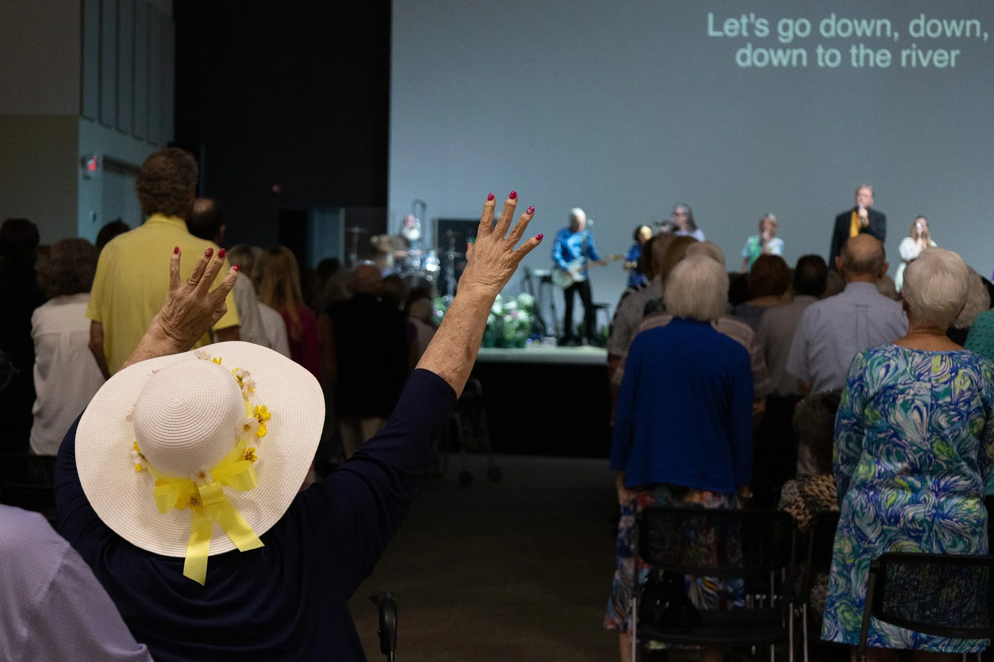 A woman in an Easter hat raises her hands in worship as the band leads a song.