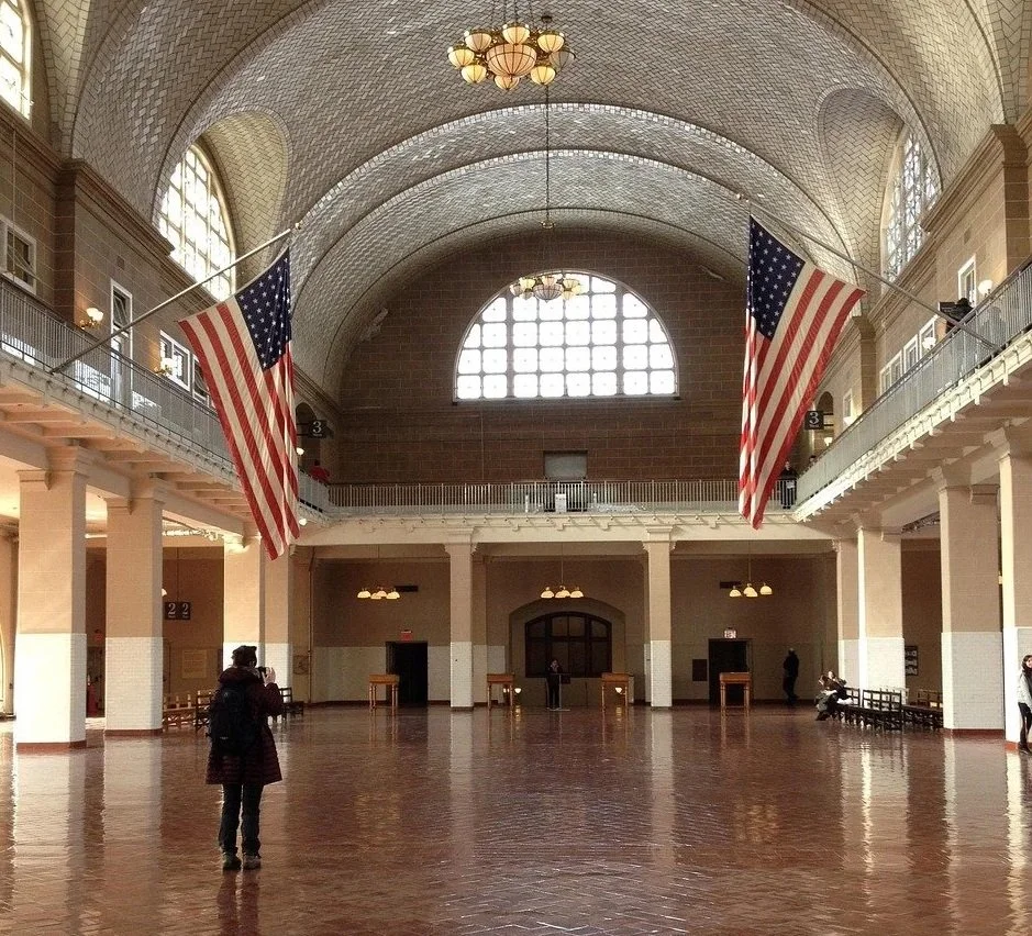 Interior of Ellis Island immigration station with a high arched brick ceiling; glowing chandeliers; American flags hanging from the balconies, and a shiny brick floor.