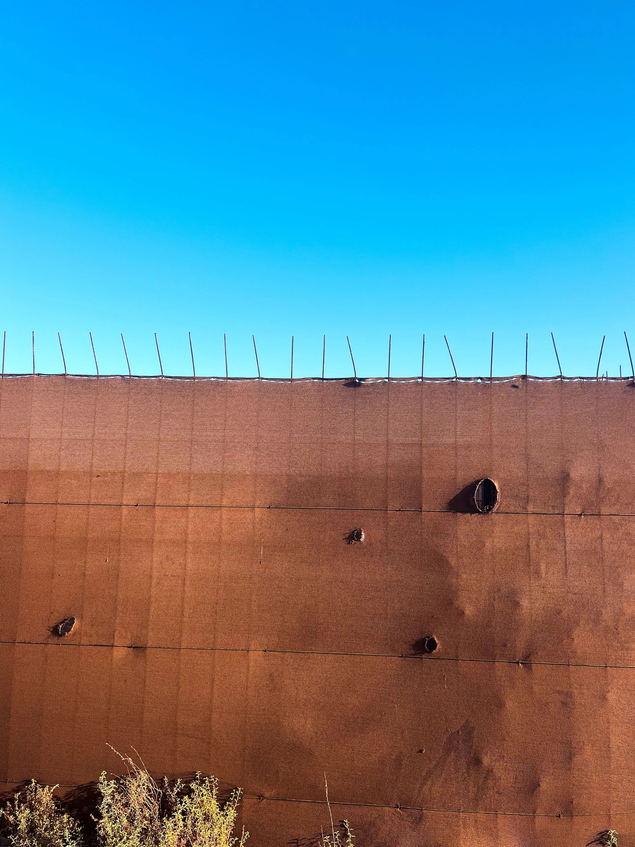 Close-up of a weathered brown stucco wall with exposed rebar along the top, holes, and shrubs below against a bright blue sky