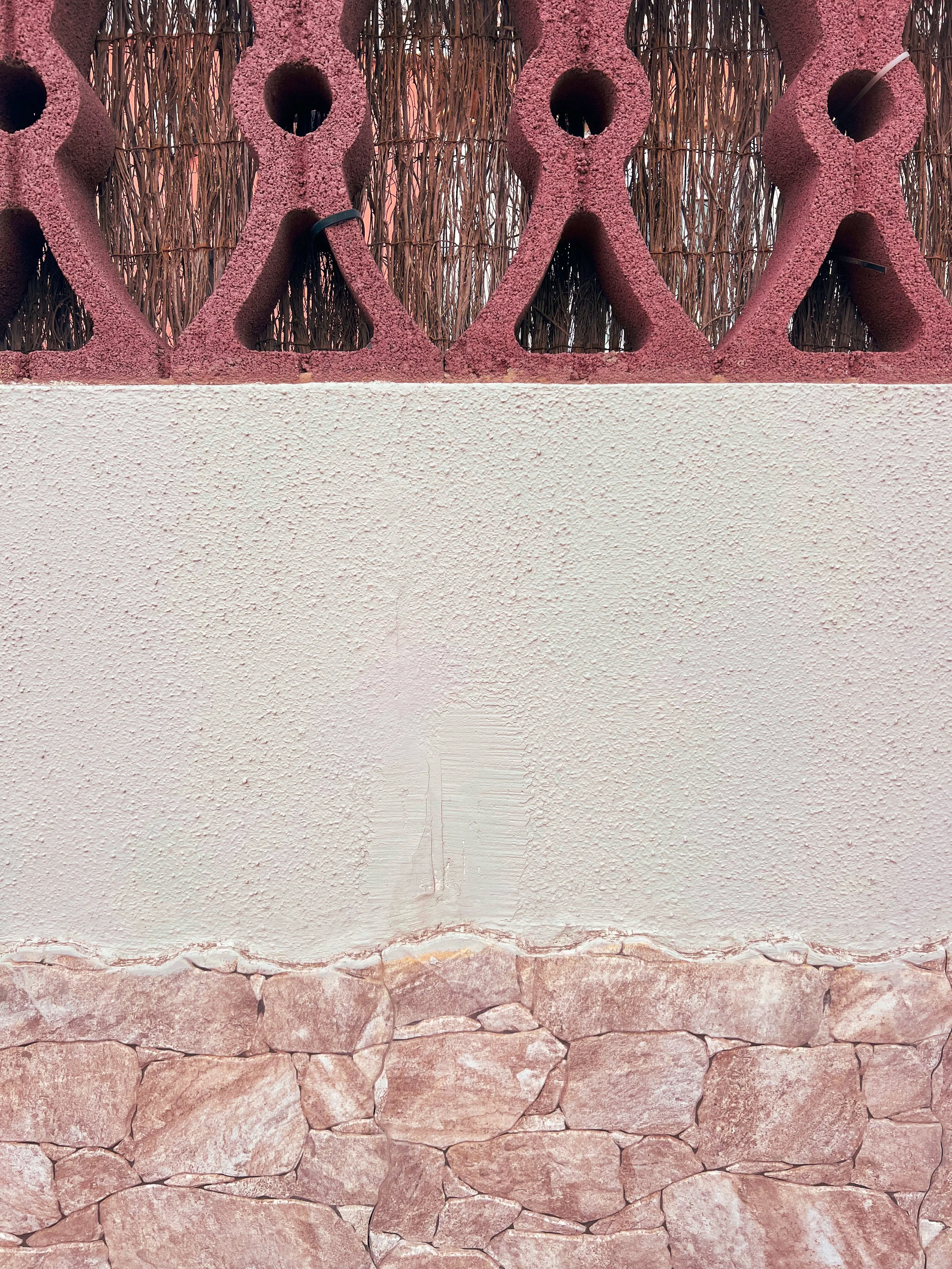 Close-up of pink textured geometric sculpture with circular openings in front of a woven reed fence and stone wall