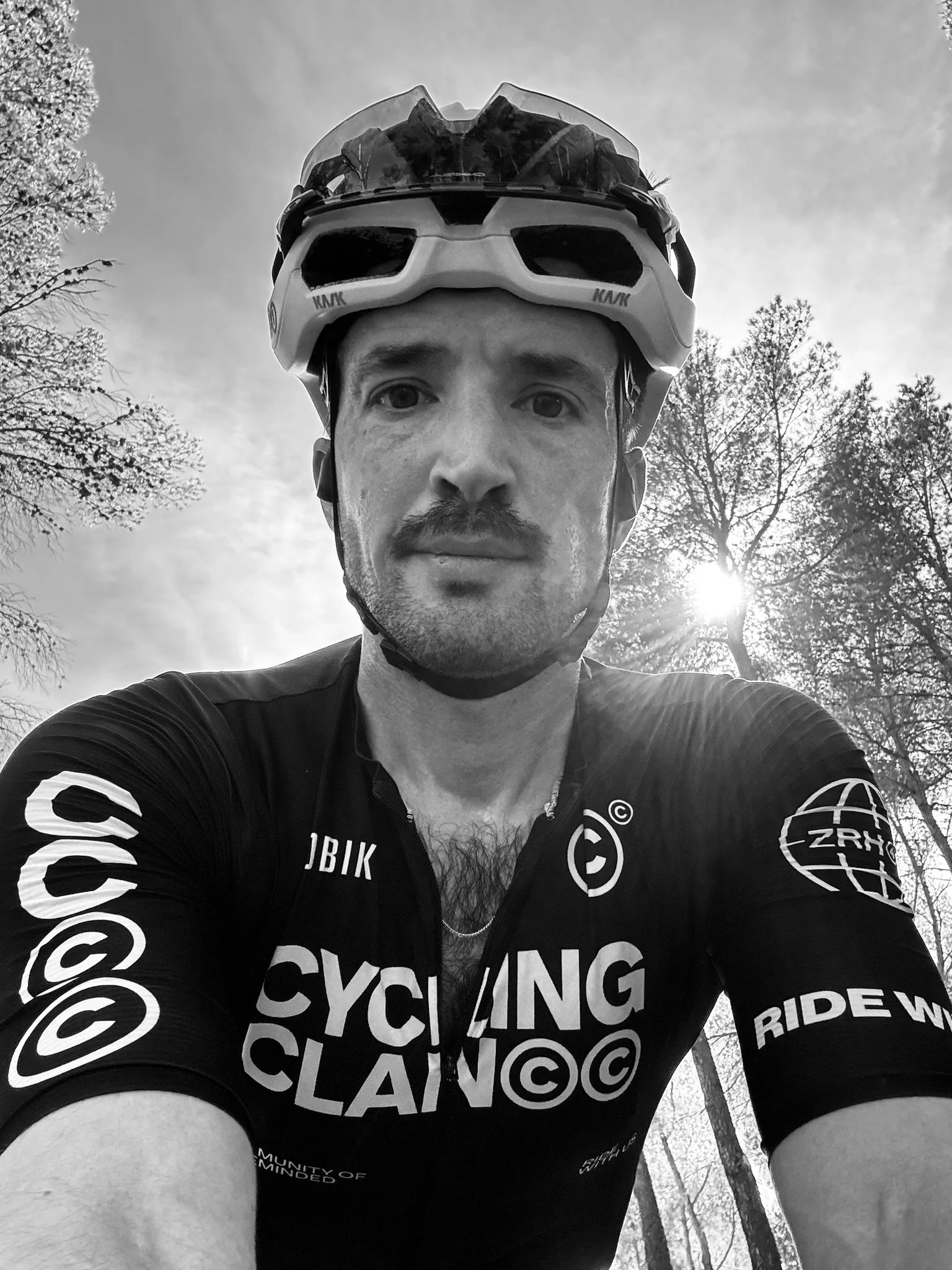 A man wearing a cycling helmet and cycling jersey, taking a selfie outdoors with trees and the sun behind him in the background.