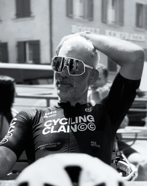 Man wearing cycling jersey and sunglasses, shading his eyes with his hand, outdoors in an urban setting.