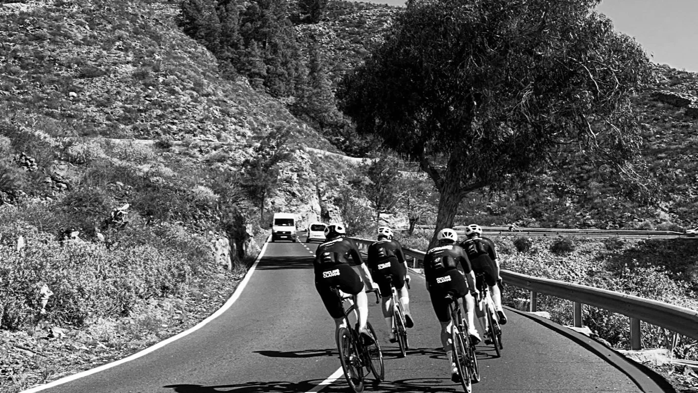 Four cyclists wearing helmets riding on a winding mountain road with a guardrail on the right, surrounded by rocky terrain and sparse trees, with a few parked vehicles in the background.