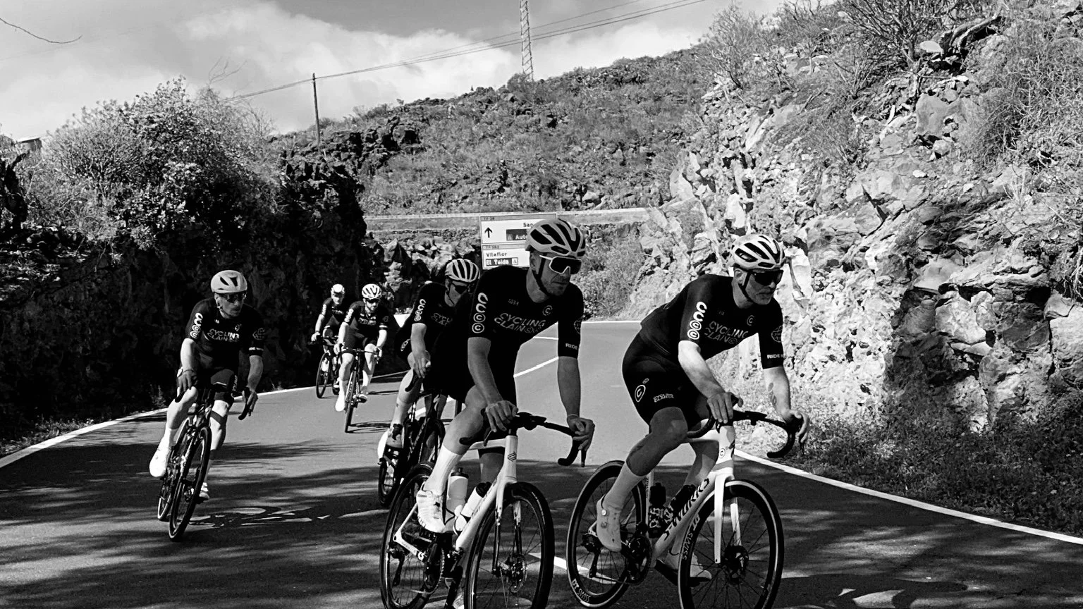 A group of six cyclists riding on a mountain road surrounded by rocky terrain and sparse vegetation, with a road sign in the background.