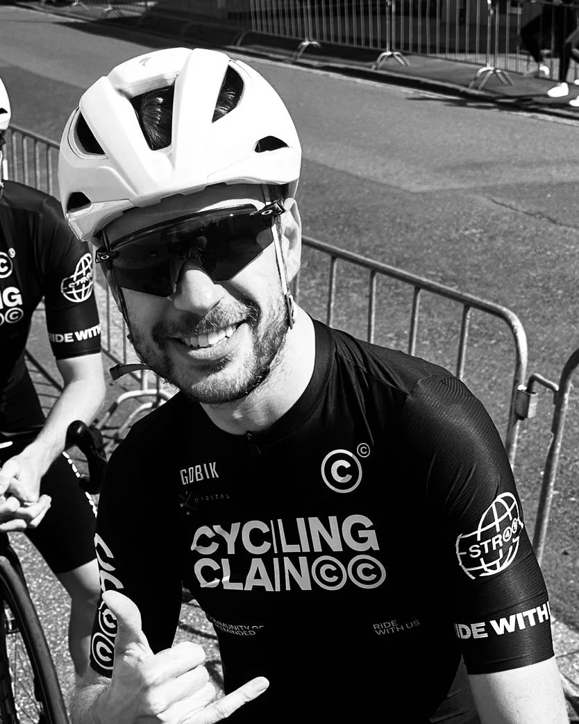 Smiling man in cycling gear giving a thumbs up, wearing a helmet and sunglasses, standing near a bike and metal barriers on a street.