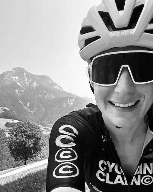 A cyclist taking a selfie outdoors, wearing a helmet and sunglasses, with mountains and trees in the background.