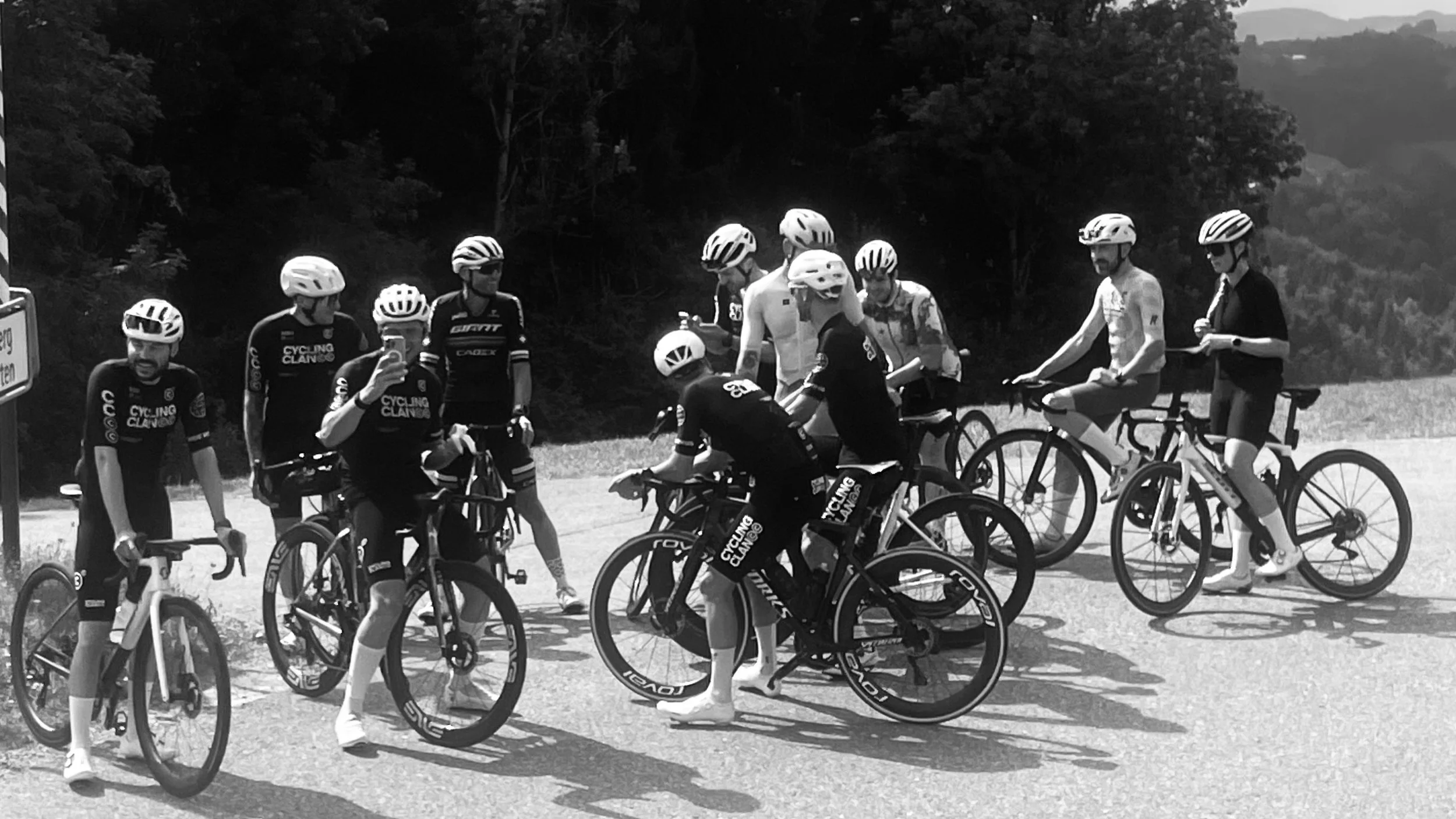 Group of cyclists wearing helmets gathered on a road, some standing with bikes and others seated on bikes, with trees in the background.