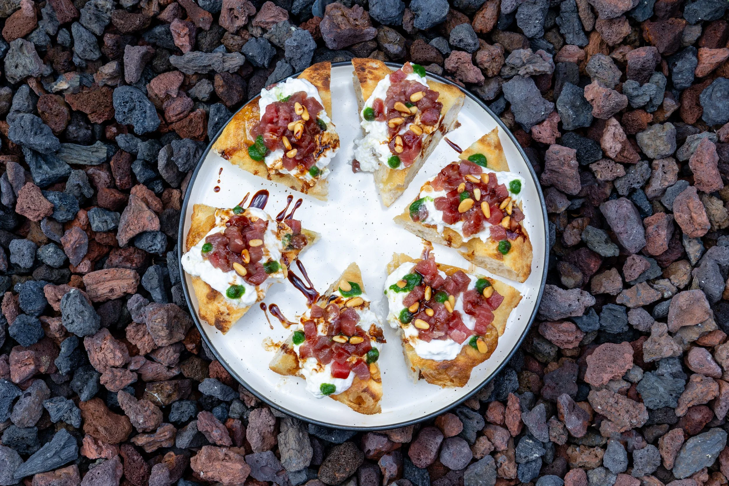 Porciones de pizza con queso, tomate, pimientos verdes, tocino, mostaza y salsa, servidas en un plato en grava negra.