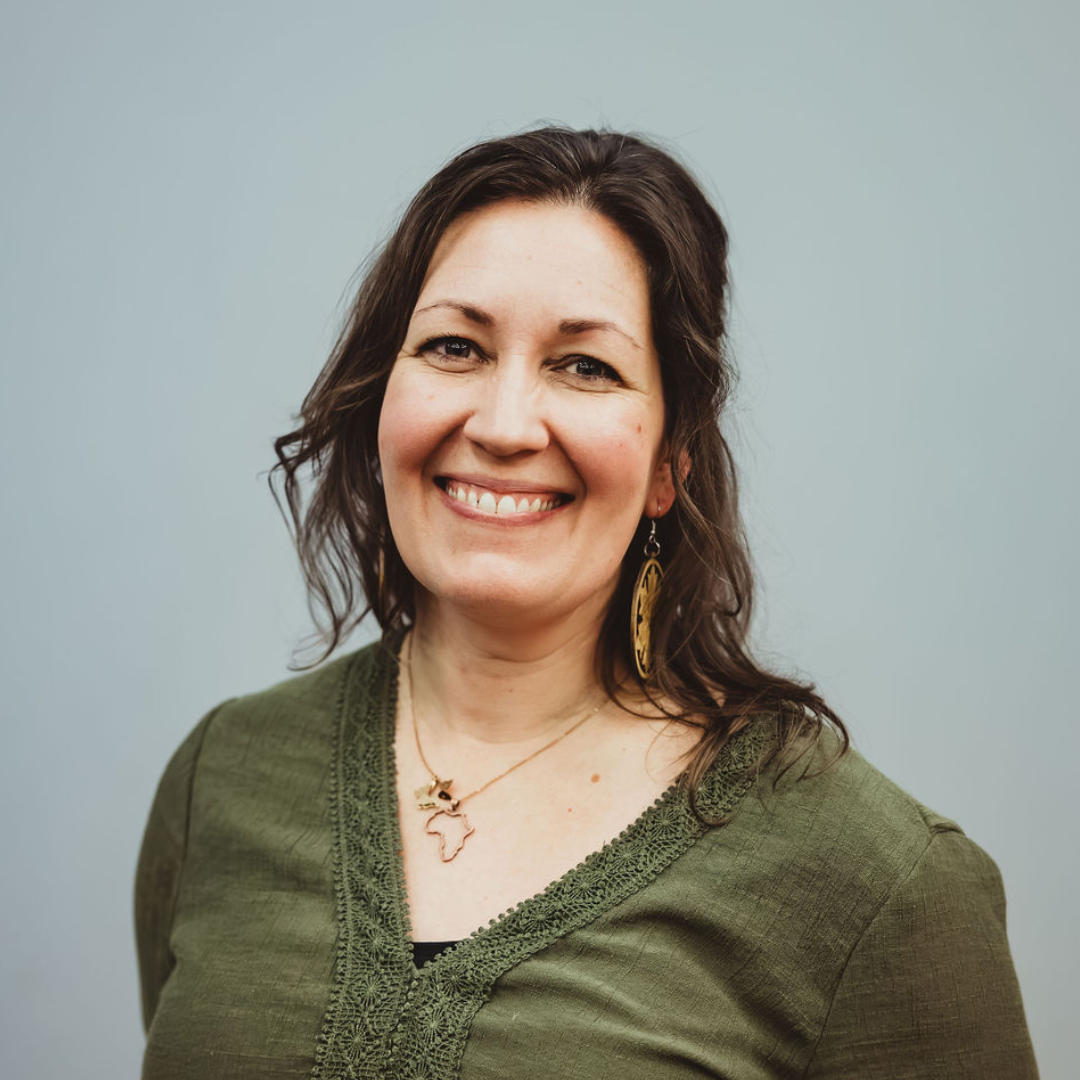 A woman with dark brown hair smiling, wearing an olive green top, earrings, and a necklace with an outline of the African continent.