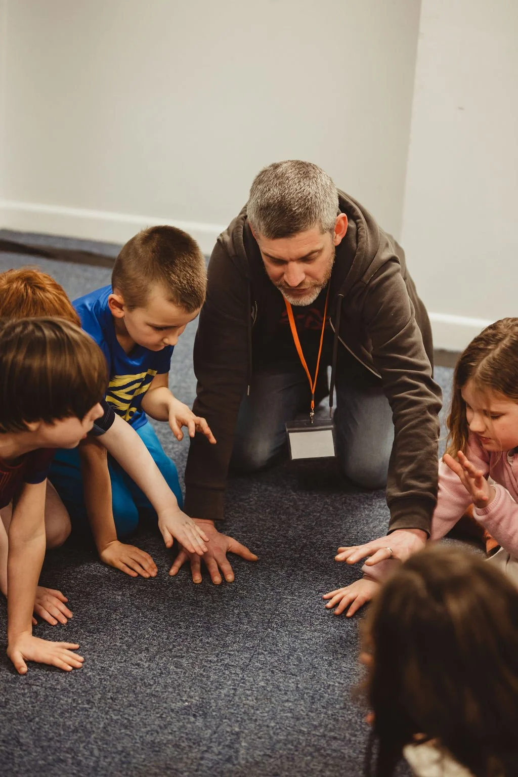 An adult man kneeling on the floor surrounded by children, all engaging in an activity together on a carpeted floor inside a room with white walls.