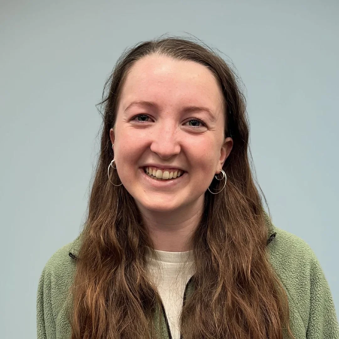 A smiling woman with long brown hair, wearing earrings and a green fleece jacket, standing against a plain, light gray background.