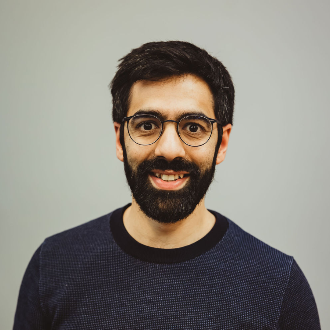 A man with dark hair, glasses, and a beard smiling in front of a plain gray background.
