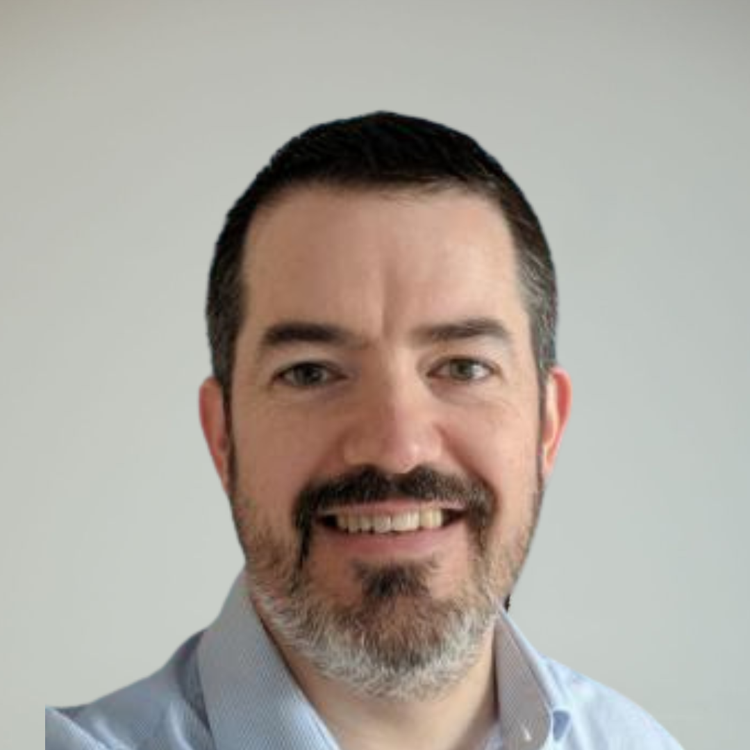 A smiling man with dark hair and a beard, wearing a light blue shirt, against a plain light gray background.
