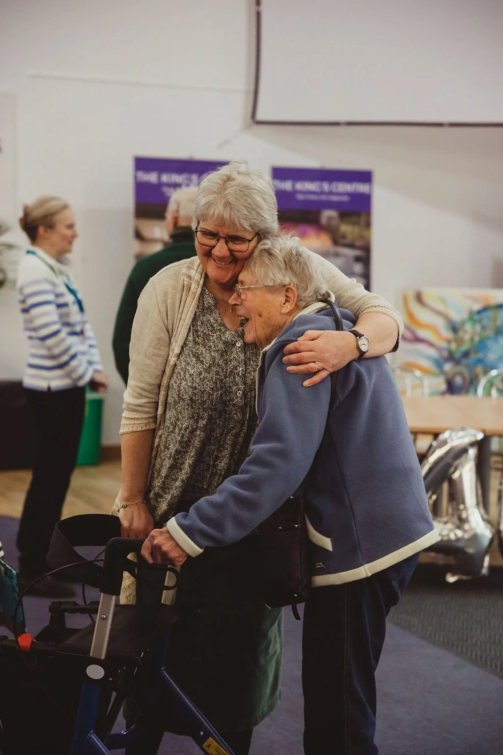 Two elderly women embrace in a warm hug at an indoor event, smiling.