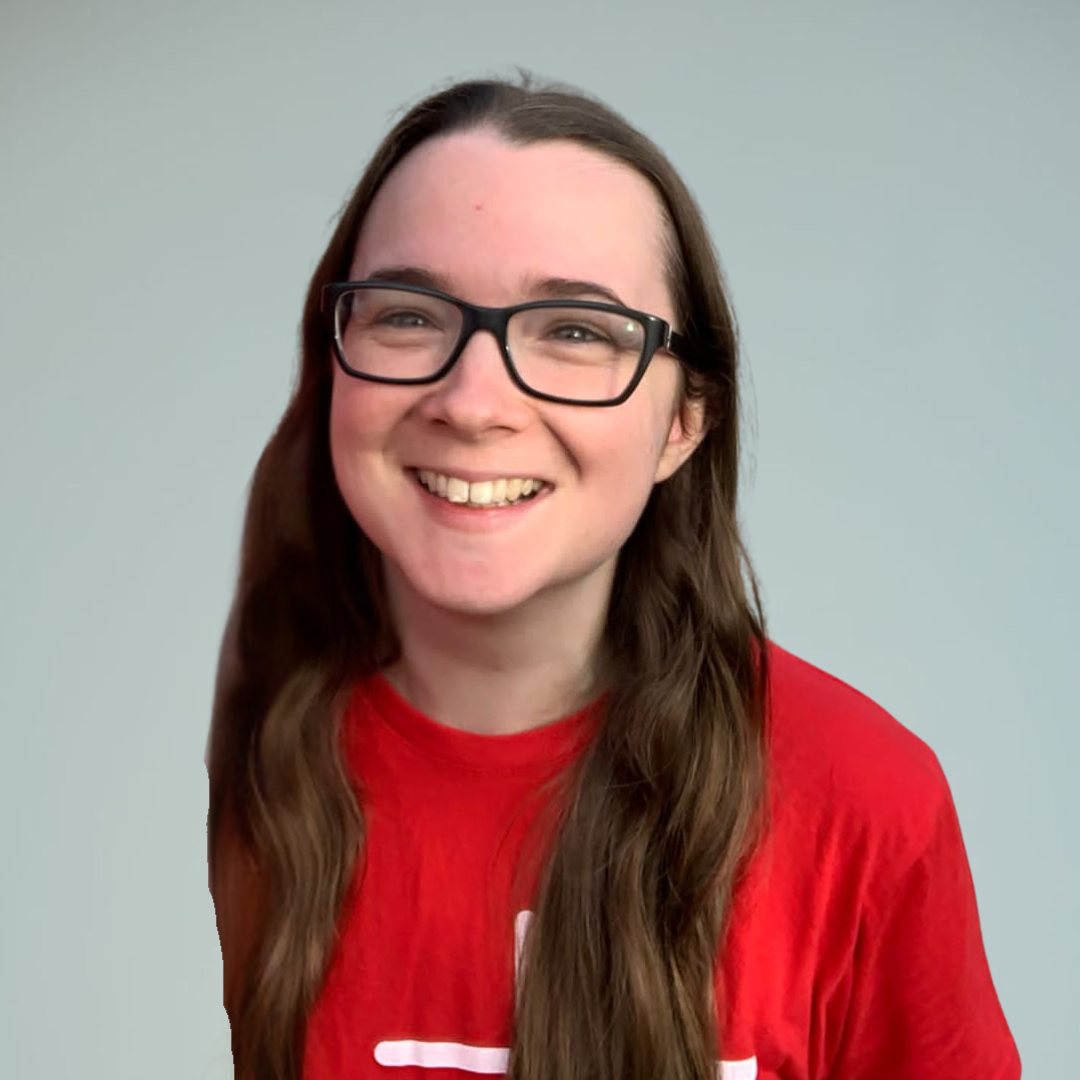 A woman with long wavy brown hair, wearing glasses and a black shirt with white star patterns, smiling in front of a plain light gray background.
