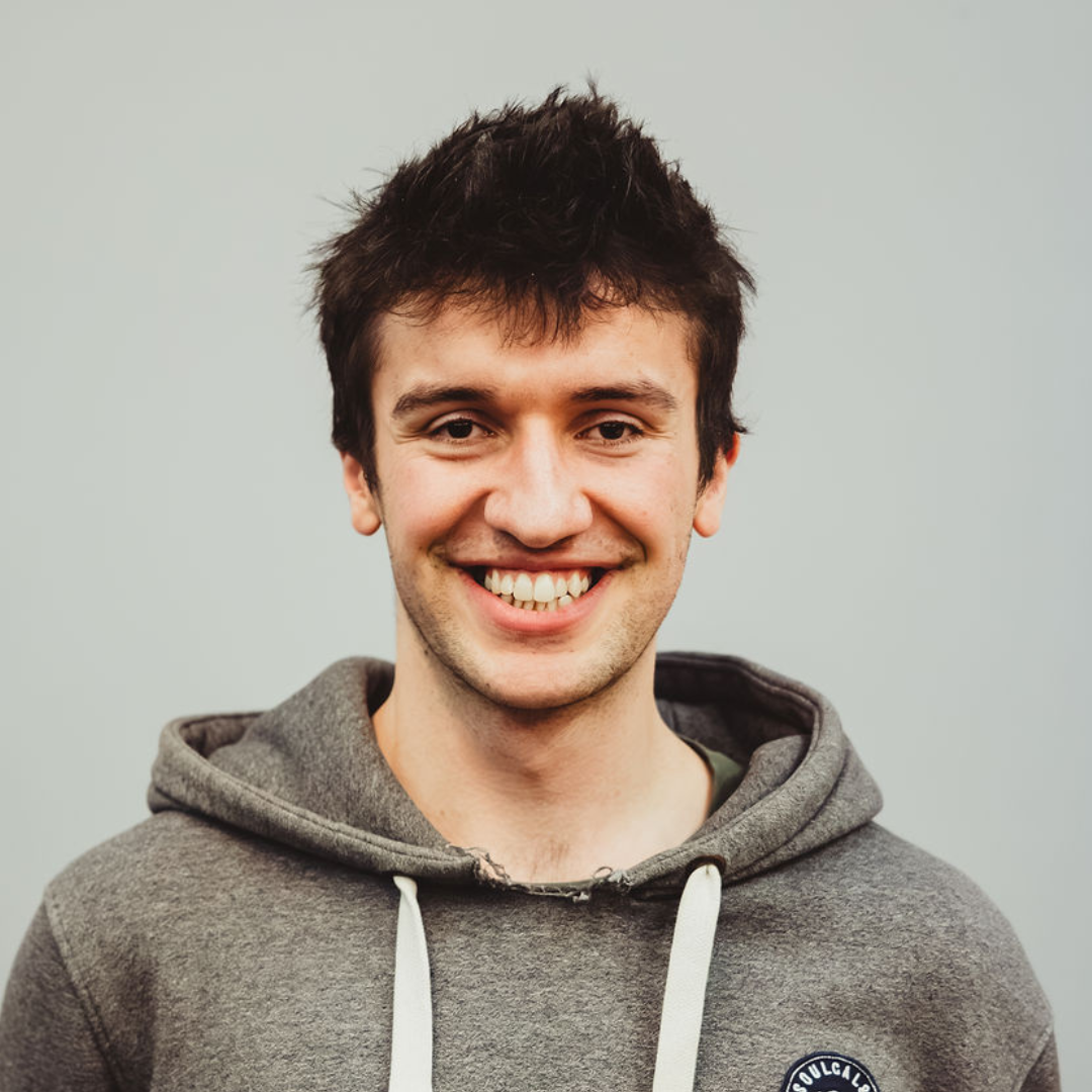 A smiling young man with dark hair wearing a gray hoodie, standing against a plain light-colored background.