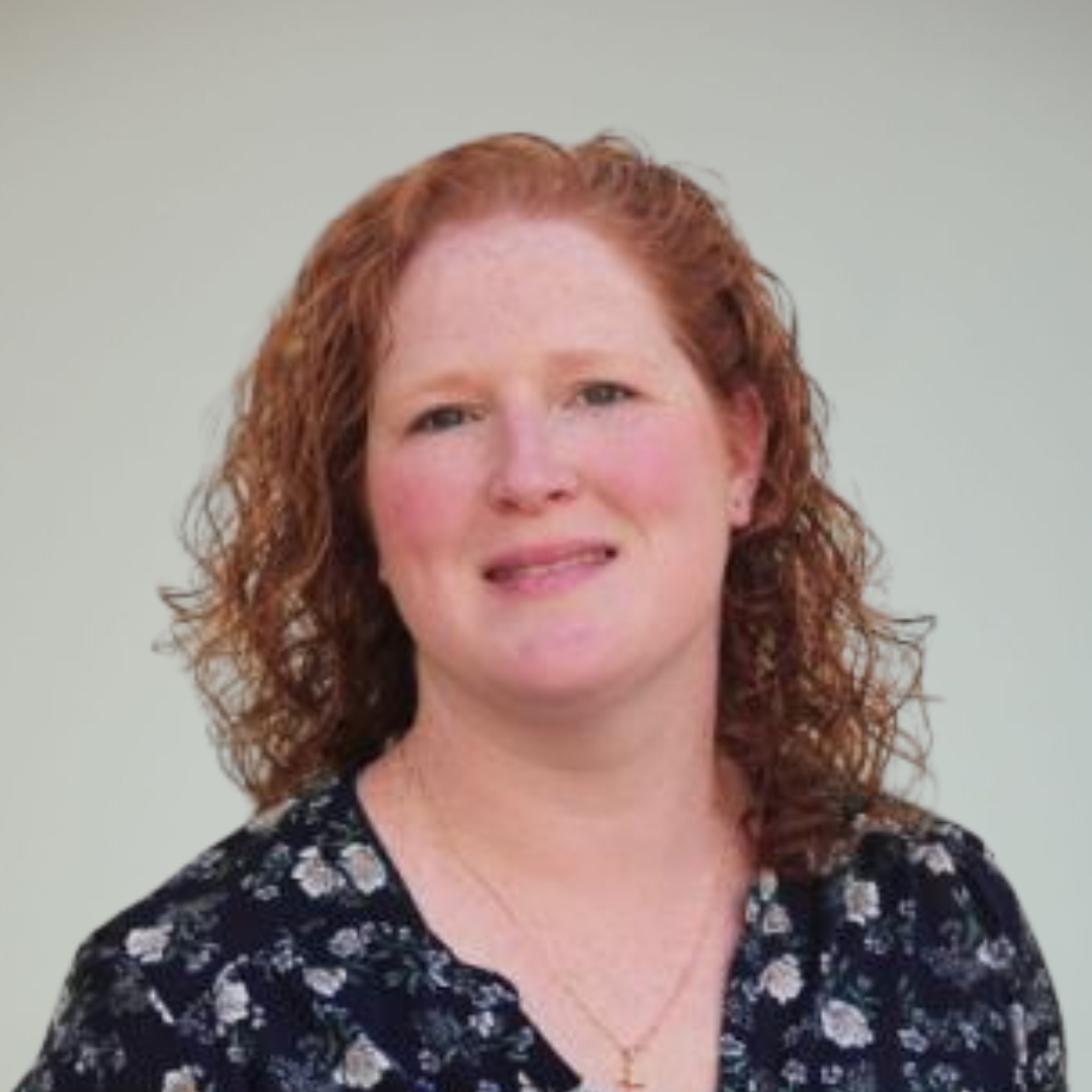 Portrait of a woman with curly red hair wearing a black top with a floral pattern, standing against a plain light background.