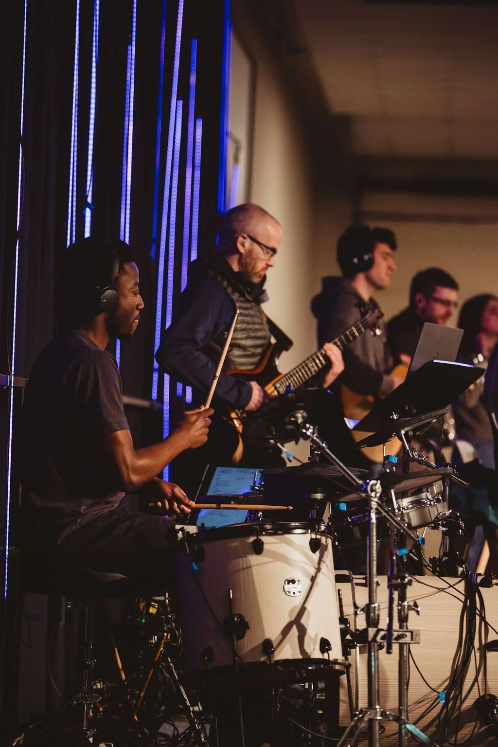 Group of musicians performing on stage with drums, guitar, and other instruments, some with headphones and music stands.