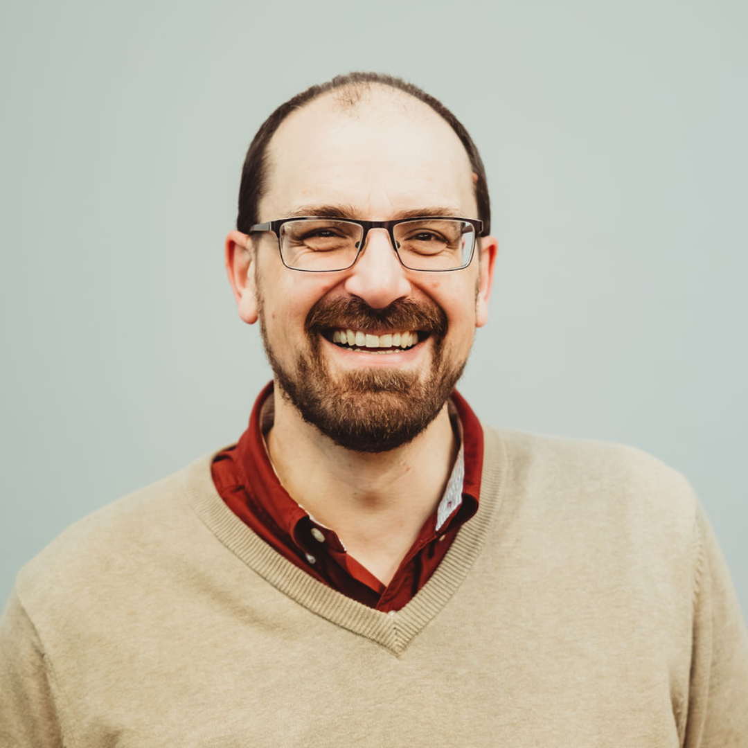 A smiling man with glasses, a beard, and a mustache, wearing a beige sweater over a red collared shirt, standing against a plain light wall background.
