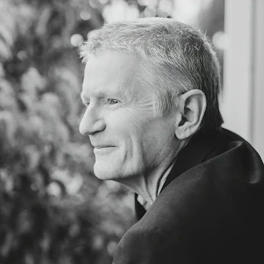 Black and white side profile of an elderly man with short gray hair, smiling gently, wearing a dark jacket. Blurred foliage in the background.