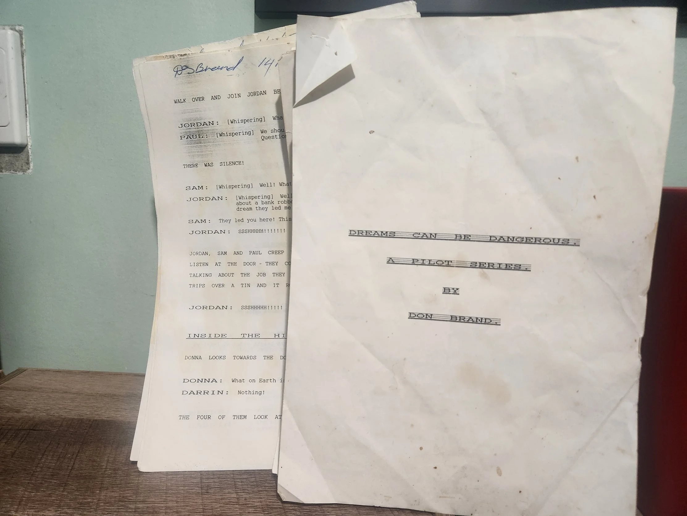 A stack of typed scripts or plays on a wooden desk, with the front page titled 'DREAMS CAN BE DANGEROUS: A PILOT SHORTS' by Don Brand. The pages behind are partially visible, showing dialogue and stage directions.