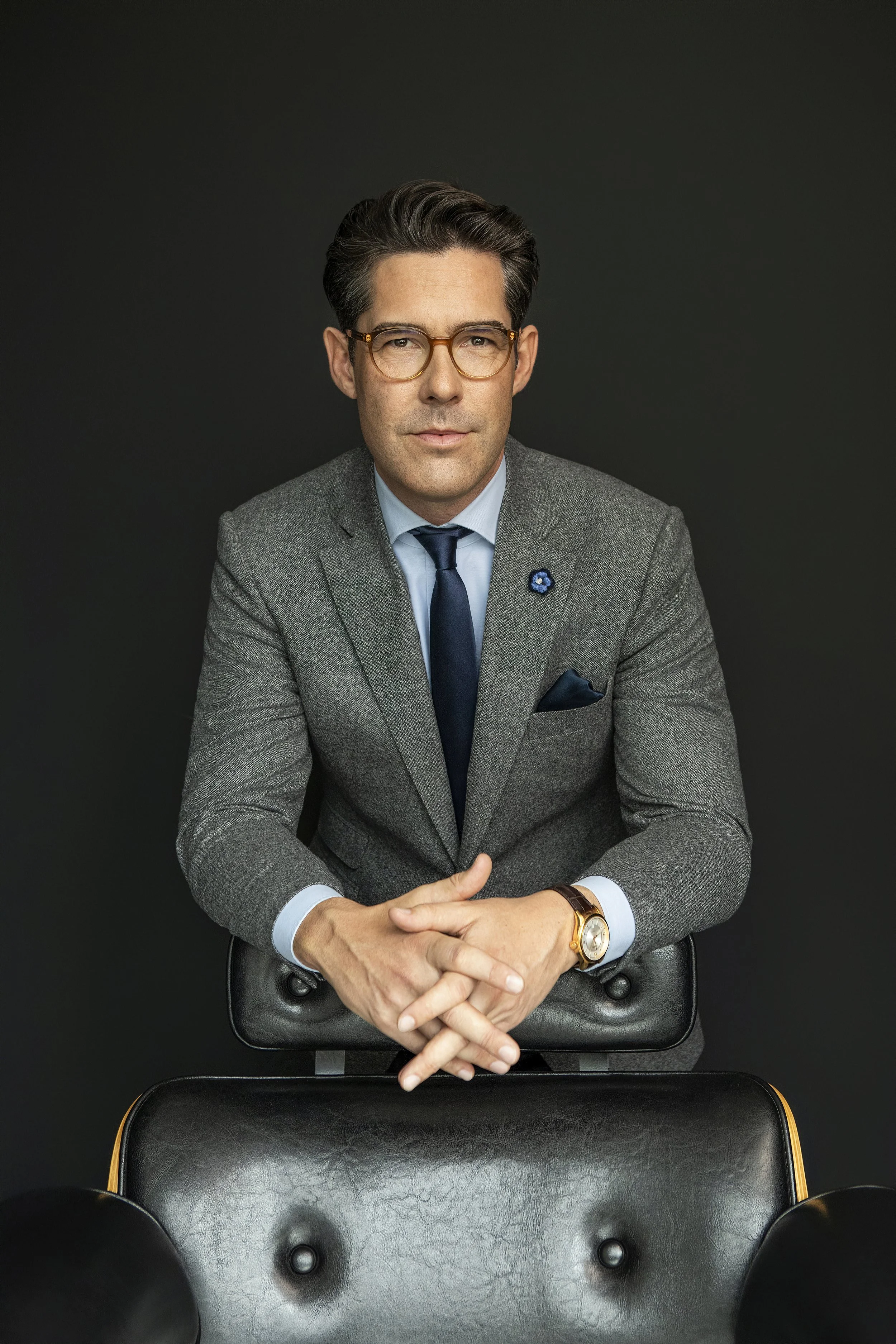 Michael Beglinger in einem grauen Anzug mit blauer Krawatte, abgestützt auf einem Sessel. Er hat dunkle, gewellte Haare, trägt Brille und eine Armbanduhr.