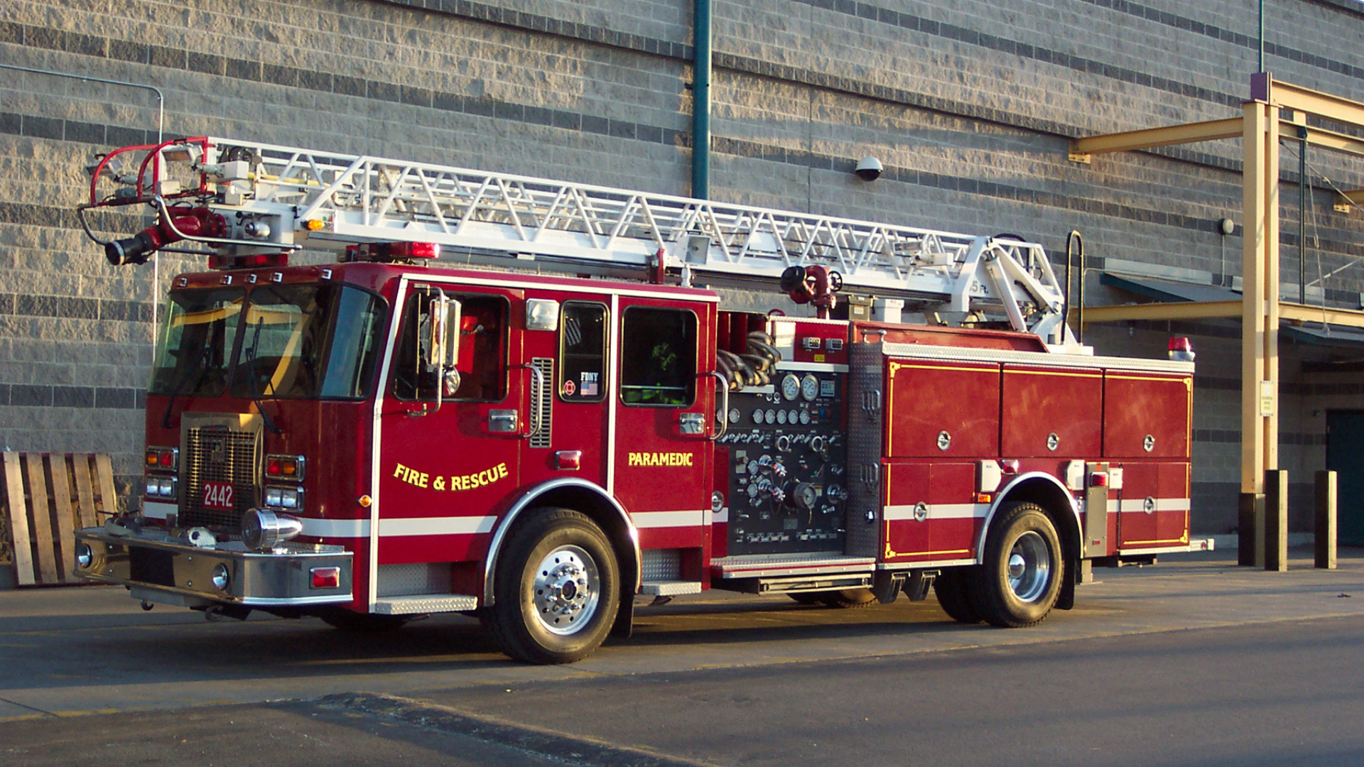 Fire truck at night with open equipment compartment, showing hoses and gauges, surrounded by firefighters.