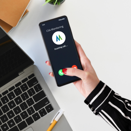 A person holding a smartphone showing an incoming call from CSA Monitoring, with a laptop and notebook on a desk.