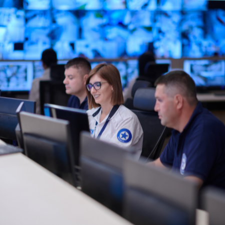 3 people working on computers in a security monitoring center with many screens displaying CCTV footage behind them.